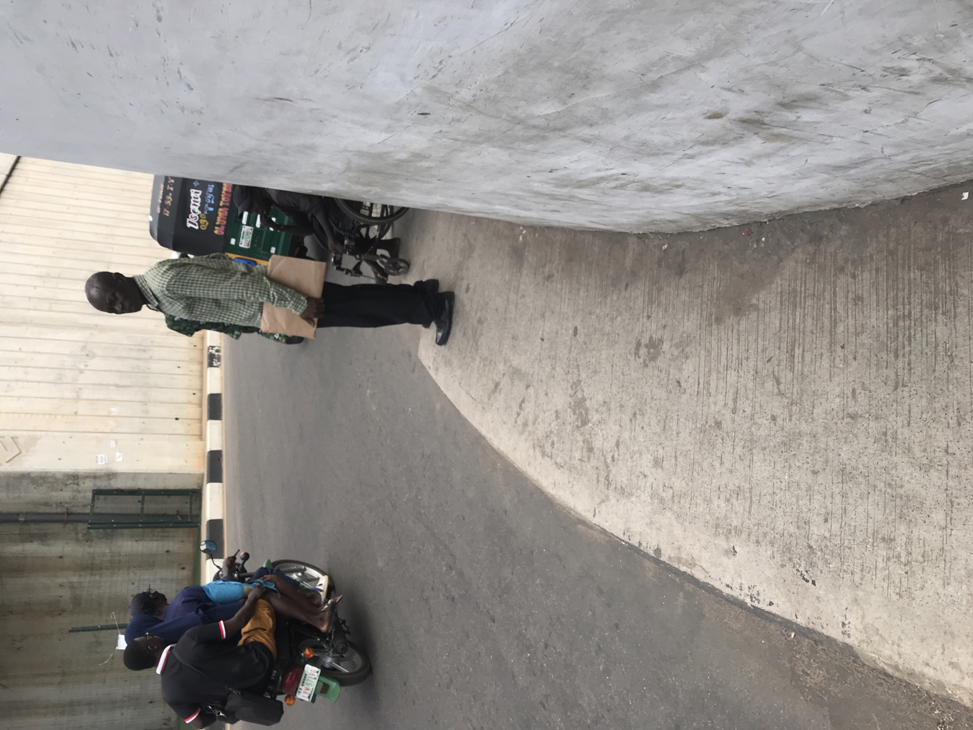 The image captures an urban scene, likely under a bridge or concrete overpass, bathed in diffuse daylight. The setting features an asphalt road curving gently on the left, bordered by a black and white striped curb that separates it from a concrete pathway and wall on the right.
Three men are visible. On the right, a man with a bald head stands on the concrete pathway, facing towards the left. He is wearing a green and white checkered long-sleeved shirt, dark trousers, and dark shoes. He holds two light brown paper bags, one in each hand, close to his body. Behind him and tucked against the concrete wall, a dark green and black tricycle taxi (keke napep) is partially visible. Text on the tricycle reads "OGUN STATE" and "OLORUN TOBI".
On the asphalt road to the left, two other men are seated on a motorcycle. The man in the front wears a dark t-shirt, while the passenger behind him wears a blue shirt. They appear stationary or moving slowly. The motorcycle has a green license plate, where the text "OGUN STATE RABANA 24" is clearly visible. The overall scene suggests a typical day in a bustling urban environment.