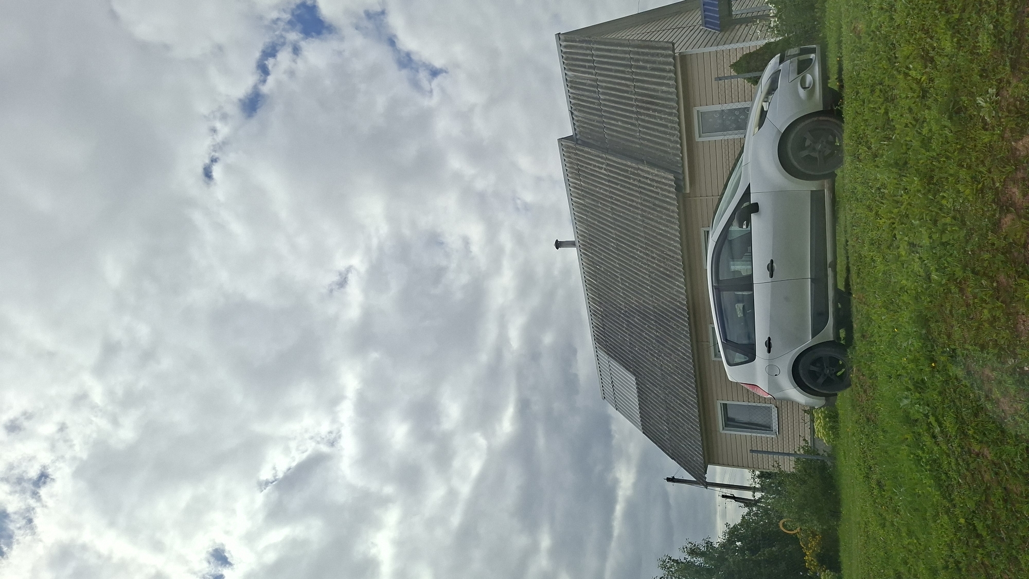 The image shows a light-colored hatchback car parked on a grassy slope in front of a light-tan, two-story house with a gray, corrugated roof. The house has several windows and a chimney. The sky is overcast with scattered blue patches. There are trees and bushes to the left of the house, and a fence post is visible. The scene appears to be in a rural or suburban area, possibly in the city of Myadzyel, Belarus, given the architectural style and vegetation. The overall impression is of a quiet, everyday scene.