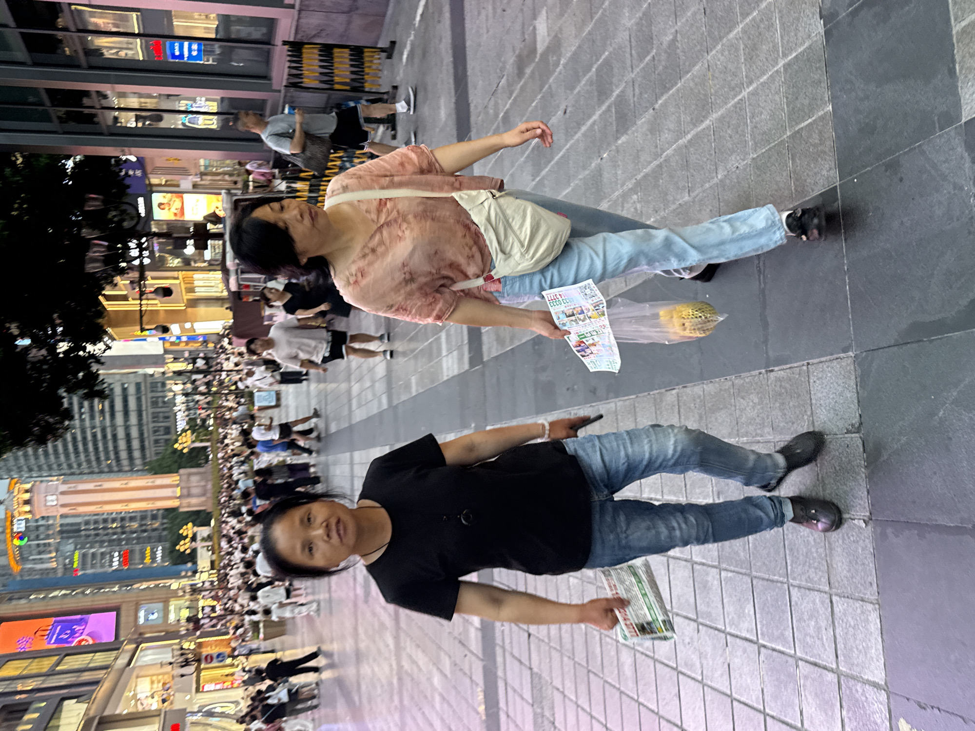 The image shows a street scene in Yuzhong District, China, with two women walking on a paved sidewalk. The woman in the foreground on the left is wearing a black t-shirt and blue jeans, holding a folded newspaper. The woman on the right is wearing a pink floral blouse and light blue jeans, carrying a beige crossbody bag and a plastic bag containing what appears to be food items. They are walking amidst a crowd of people, and buildings with illuminated advertisements are visible in the background. The street is lined with shops and commercial establishments. The lighting suggests it is either dusk or a brightly lit urban environment.