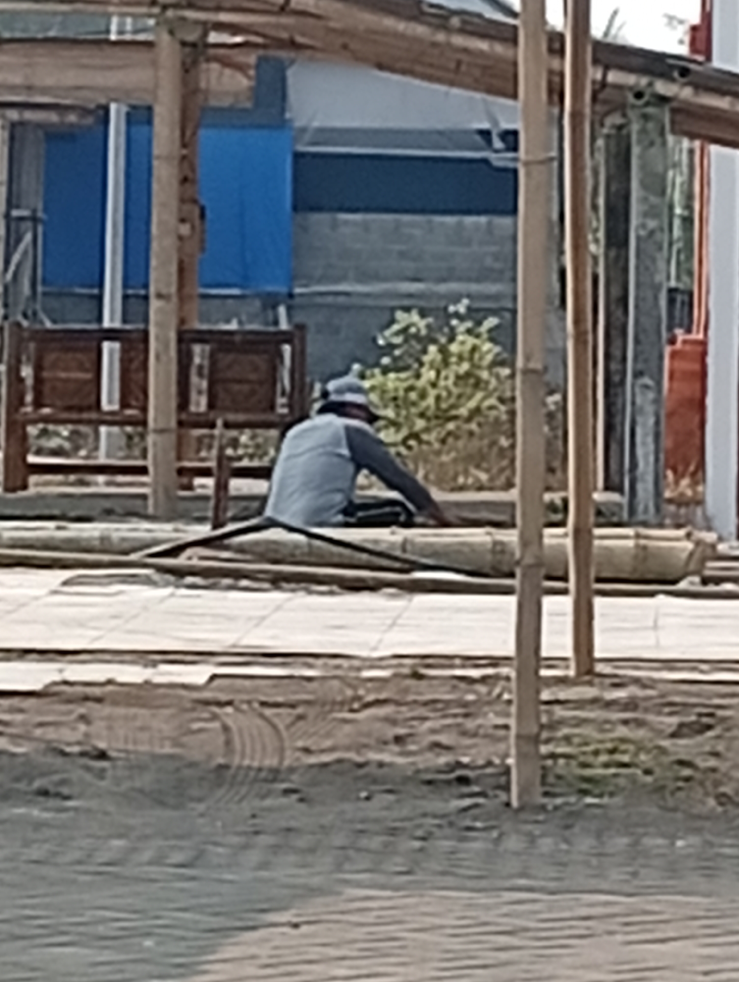A person wearing a blue cap and light blue long-sleeved shirt is sitting on the ground in front of a structure with wooden poles and beams. The person is partially obscured by bamboo poles arranged in front of them. In the background, there is a building with blue panels and grey brickwork. Two dark wooden benches are visible on the left. The ground in the foreground is paved with grey interlocking bricks. This scene is located in Srandakan, Indonesia.