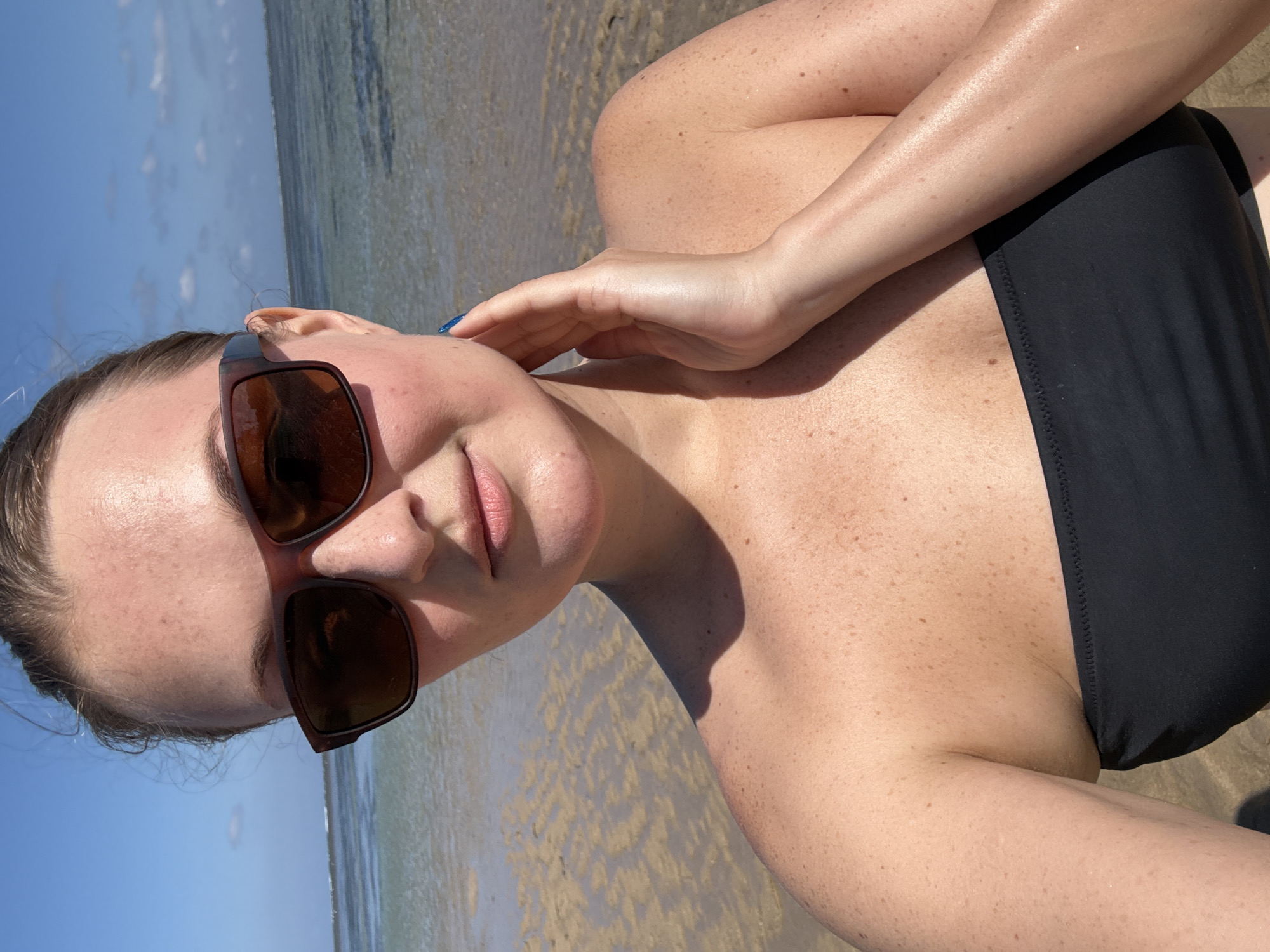 A young woman with fair skin and freckles is posing for a selfie on a beach. She is wearing a black strapless bikini top and brown sunglasses. Her right hand is touching her neck. The background shows a shallow body of water with sandy seabed and a clear blue sky. The location is identified as Denpasar, Indonesia.