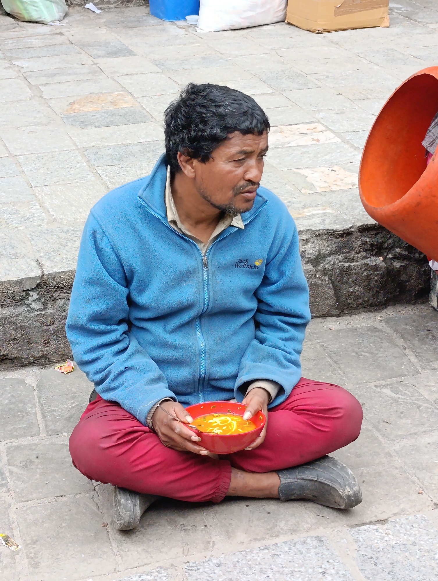 A man with dark, curly hair and a light beard is sitting cross-legged on a cobblestone street, holding a red bowl of yellow food. He is wearing a blue zip-up jacket over a light-colored shirt and bright red pants. His shoes appear to be dark, worn, and possibly rubber. To his right, a large, bright orange object, possibly a container or a piece of machinery, is partially visible. The background consists of the stone pavement, some debris on the ground, and a rough stone wall. The scene appears to be outdoors, possibly in a market or public area. The man's gaze is directed to his right, away from the camera. There is no clear indication of interaction between the man and anyone else in the frame. The lighting suggests it is daytime. The overall impression is that of a candid street photograph capturing a moment of repose. The image is not specifically identifiable as being in Manali, India, based solely on its visual content.
