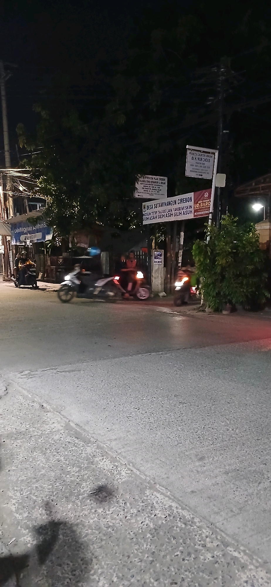 This is a nighttime outdoor photograph of a street in Sutawinangun, Indonesia. The scene depicts a road with several motorcycles. One motorcycle is in motion in the foreground, with its headlights on and taillights illuminated. Another motorcycle is parked on the left side of the image, and two other motorcycles are visible further down the street. Two individuals are seated on one of the moving motorcycles, and another individual is standing near a parked motorcycle. Several signs are visible, including one that reads "PRAKTEK DOKTER UMUM dr. H. NUR CHOLIS" and another that says "DESA SUTAWINANGUN CIREBON". The environment appears to be a moderately lit urban or semi-urban area with trees and buildings visible in the background. The road surface is asphalt.