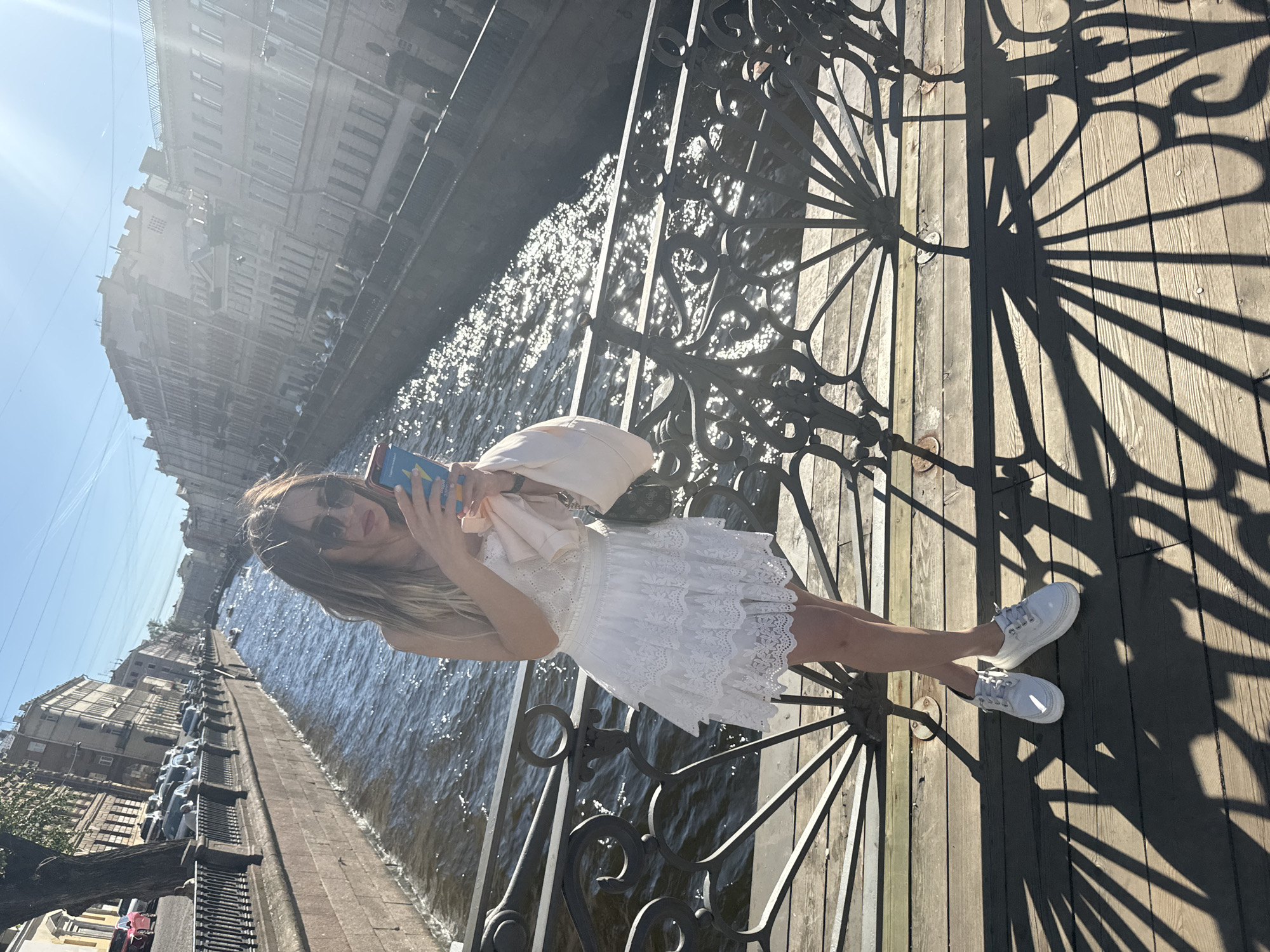 A young woman with blonde hair and sunglasses stands on a bridge in Saint Petersburg, Russia, holding a mobile phone. She is wearing a white lace dress and white sneakers. The bridge has decorative black metal railings. A canal runs alongside the bridge, with buildings and trees on the other side. The sun is shining, casting shadows on the wooden planks of the bridge.