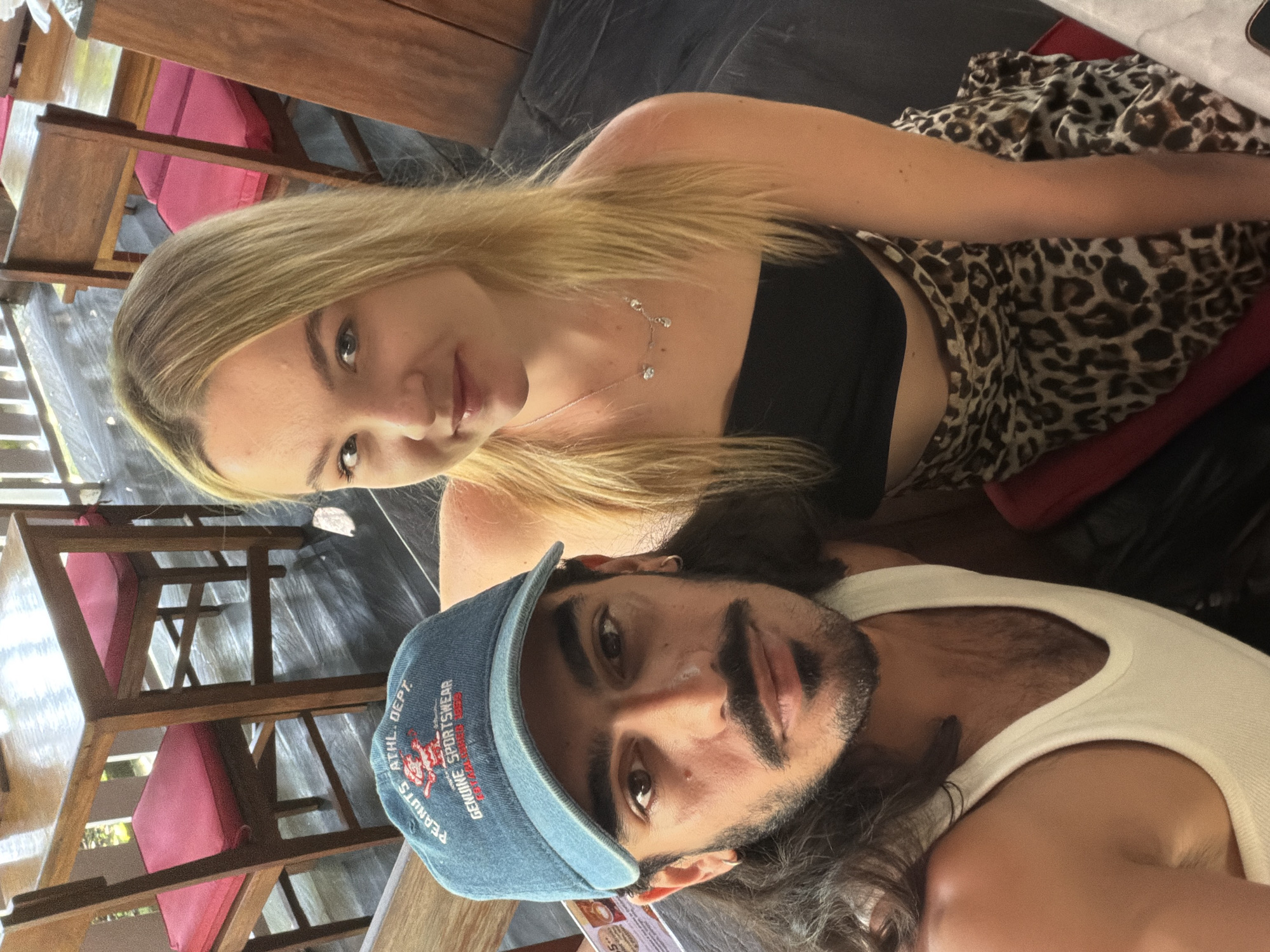 A woman with blonde hair and a black top and leopard-print skirt is posing with a man with long hair and a baseball cap. They are sitting at a wooden table with wooden chairs and pink cushions. The location is North Kuta, Indonesia.
