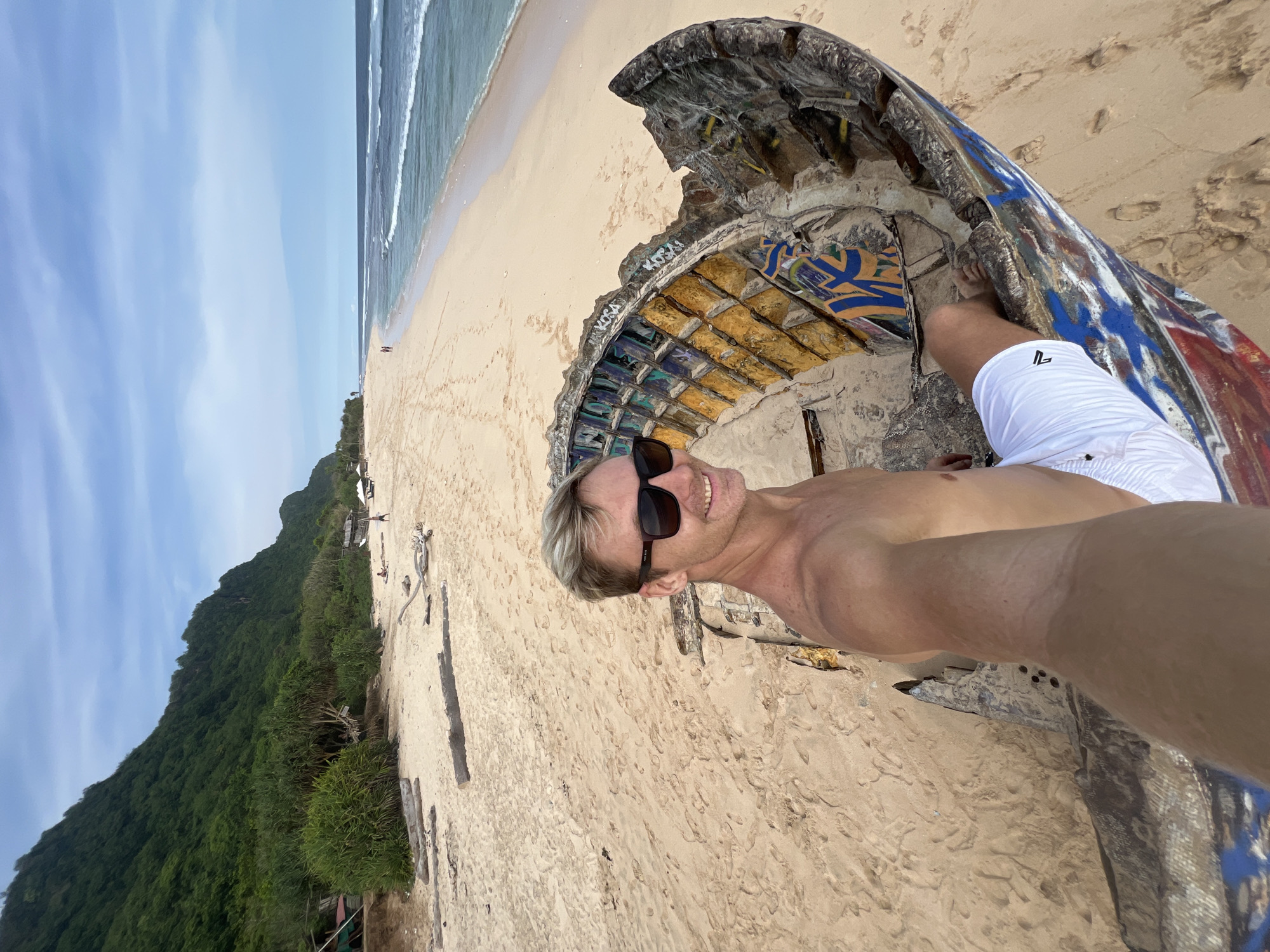 On a sandy beach in Pecatu, Indonesia, a man is posing in front of the fuselage of an airplane that has been partially destroyed. The plane’s interior is decorated with graffiti and remnants of its original structure. The man is fair-skinned, blond, wearing sunglasses, and shirtless with white shorts. He is positioned within the plane’s structure. The beach extends into the background, where a body of water meets the shore and a lush, green landscape rises to the left. The sky is partially covered with white clouds. Scattered debris is noticeable on the beach.