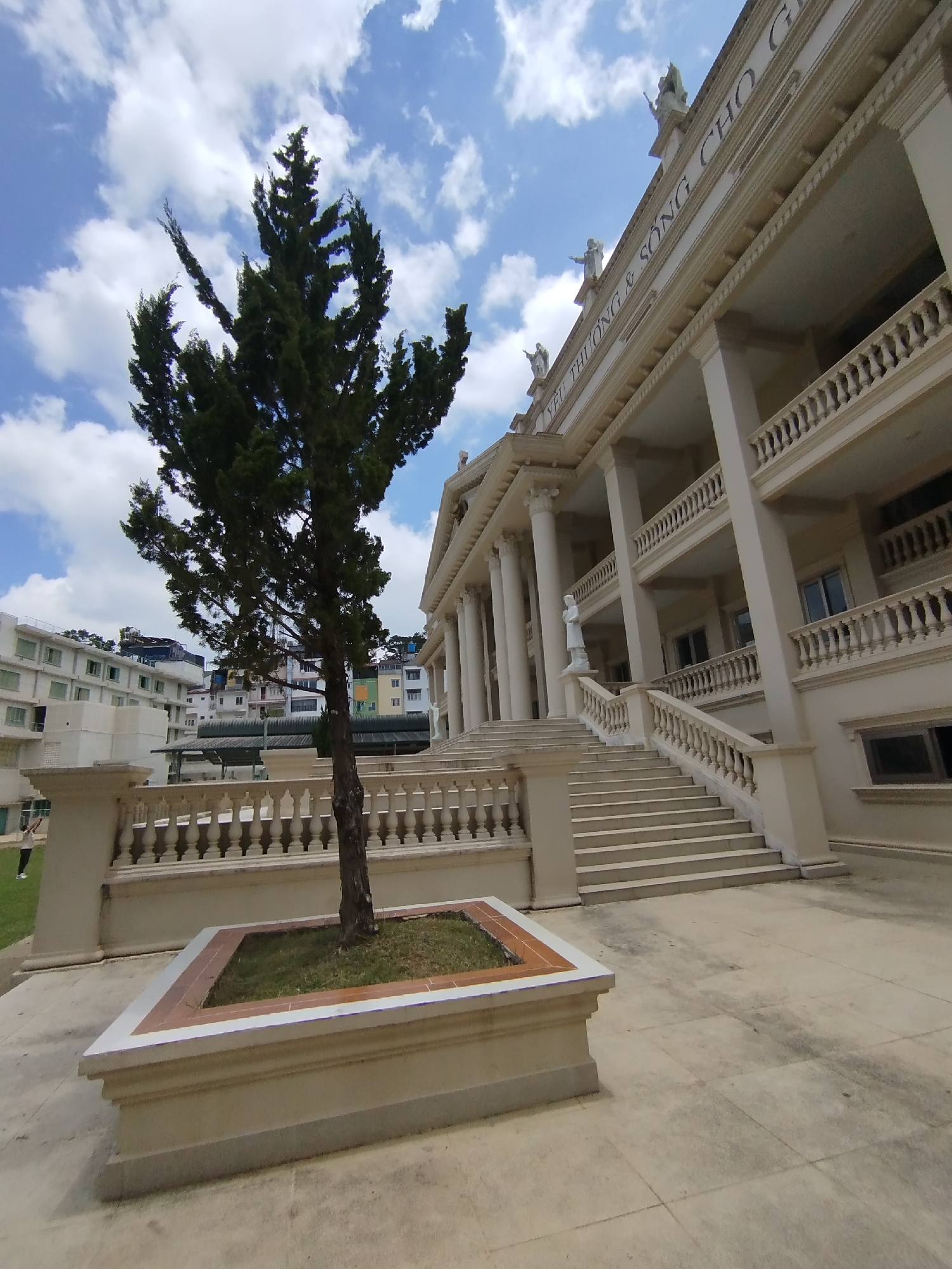 A low-angle shot shows a stone building with classical architectural elements in Da Lat, Vietnam. The building has a pale beige facade, adorned with columns and statues. A wide staircase leads up to the main entrance. In front of the building stands a rectangular planter containing a slender, dark green tree with an irregular crown. The sky above is blue with scattered white clouds. Other buildings can be seen in the background. A section of grass with a person standing on it appears on the left edge of the image. The building's upper facade has writing on it.