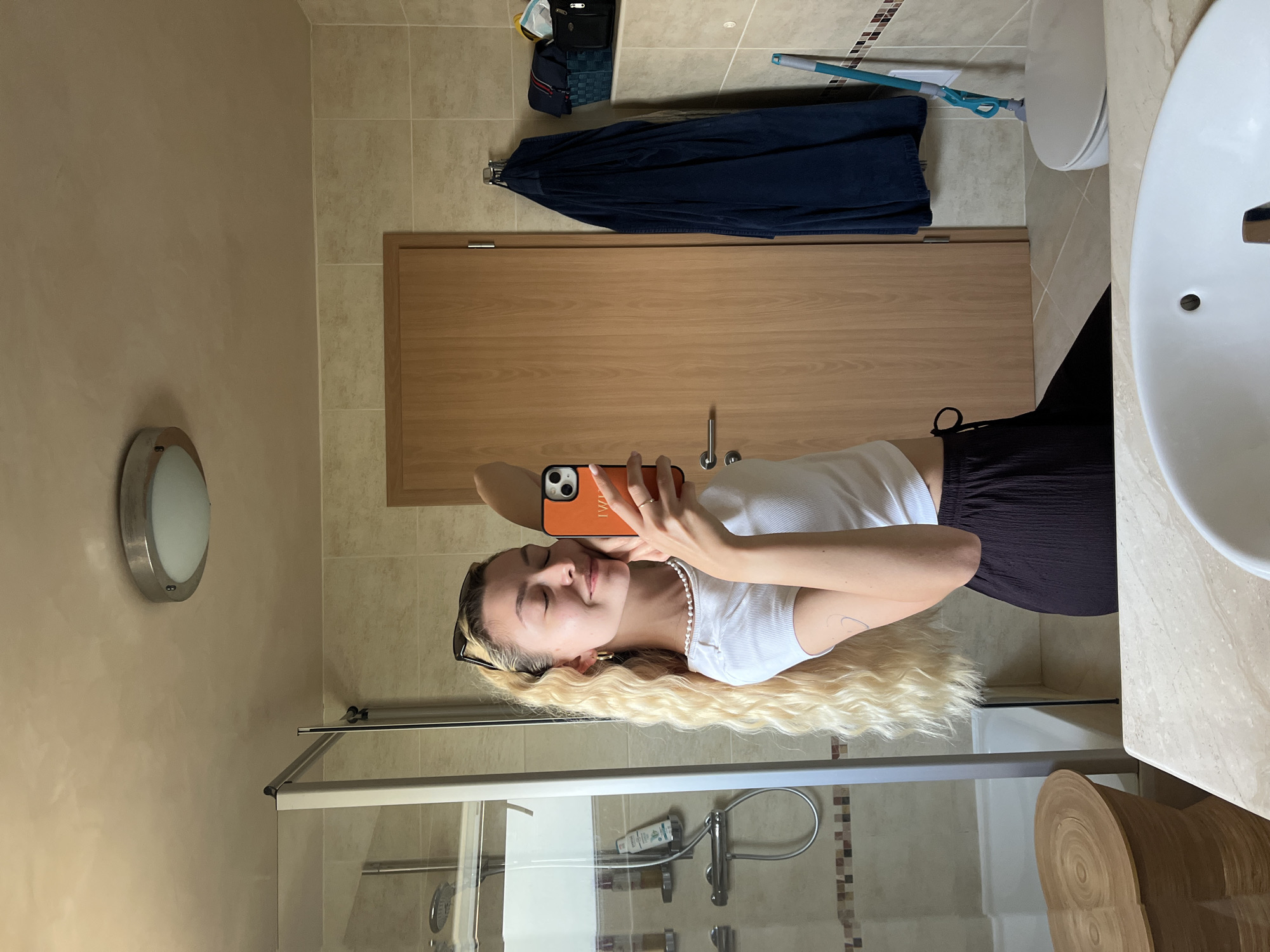 A young woman is captured in a selfie taken in a well-lit modern bathroom, with the image rotated 90 degrees clockwise from its intended upright orientation. She has long, wavy blonde hair flowing down, and is wearing a white ribbed t-shirt and dark pleated pants. A pearl necklace adorns her neck, and sunglasses are pushed up onto her head. She holds an orange-cased smartphone, with the word 'LUAI' visible on the case, and a gold-colored ring on her finger. Her eyes are closed, and a gentle smile plays on her lips, conveying a relaxed and content mood.
The bathroom features light beige tiled walls and floor, with a decorative mosaic tile strip running along one wall near the shower. One wall is a plain light tan color with a textured finish. Visible elements include a wooden bathroom door, a white sink, and a toilet. A glass-enclosed shower cabin is also present, reflecting bottles of toiletries inside. A round, silver-trimmed light fixture is mounted on the wall.
To the left of the door, a dark blue garment hangs on a hook, and a black bag with a yellow object is placed on a shelf. A blue mop with a green handle is propped against the toilet. Near the sink, there is a small wooden stool or decorative item with a concentric circular pattern. The overall impression is one of a clean and functional bathroom environment, likely during daytime.