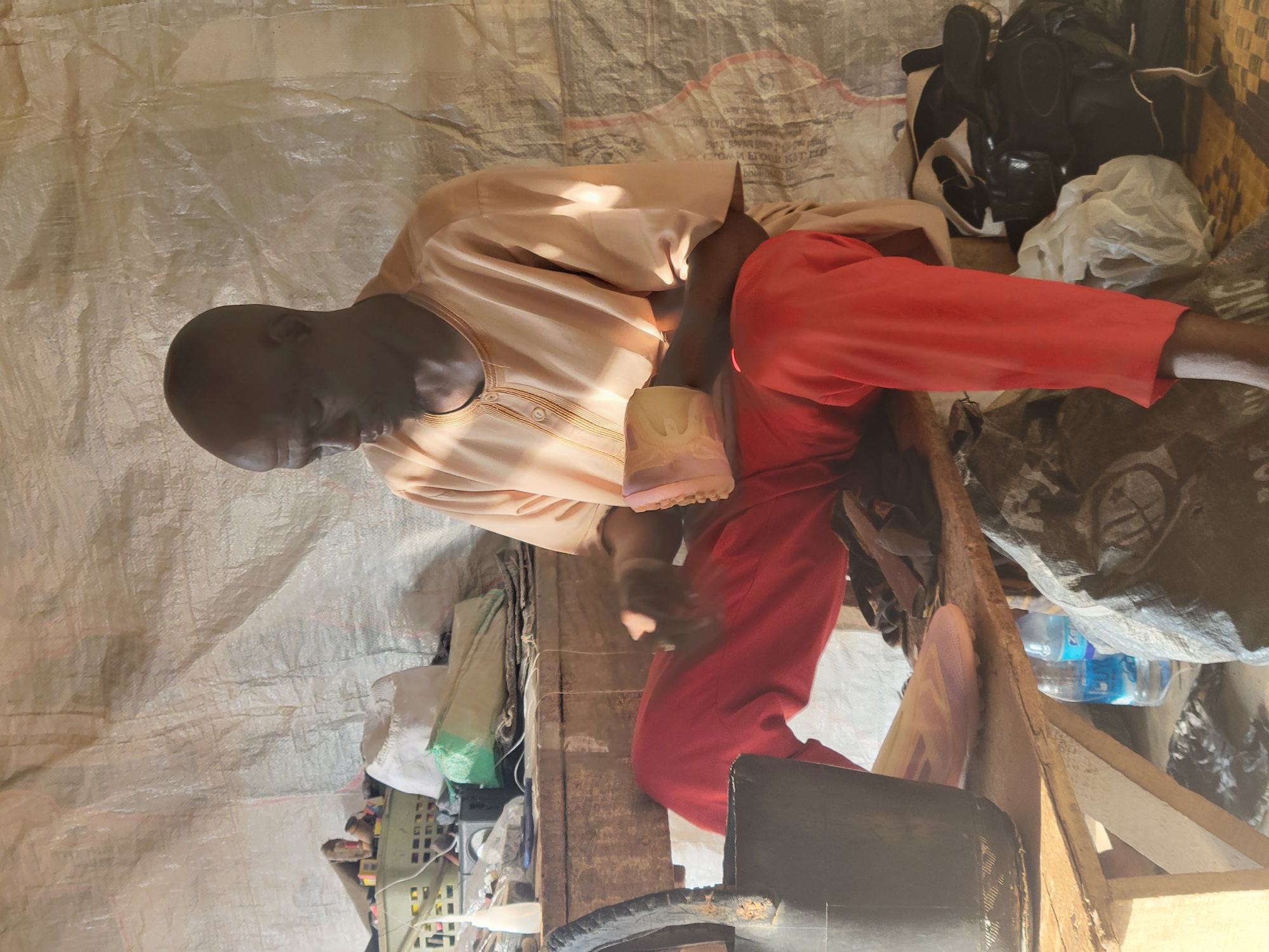 In a makeshift outdoor workshop in Madalla, Nigeria, a bald, dark-skinned man wearing a light-colored, collared tunic and bright red pants is seated on a wooden bench. He is holding a light pink object, possibly a shoe or a part of one, in his hands, appearing to be working on it. To his left, a black bucket rests on the ground. The background consists of a large, translucent tarp stretched taut, with various items like bags, bottles, and tools visible on shelves and the ground. Sunlight casts shadows, indicating an outdoor setting.