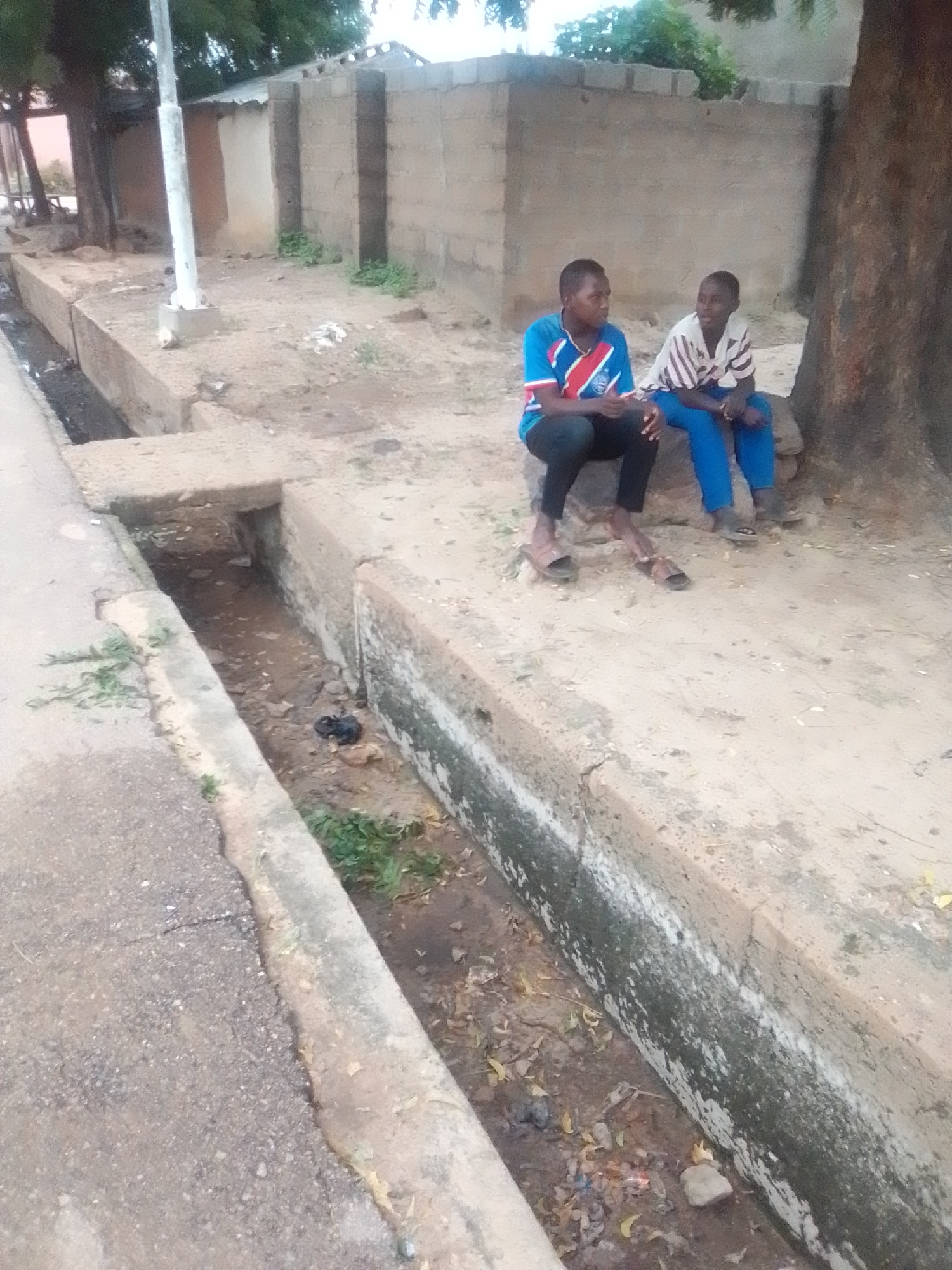 Two young males are seated on a concrete ledge next to a dry drainage ditch. The individual on the left wears a blue, red, and white striped t-shirt and black trousers. The individual on the right wears a white and striped t-shirt and blue trousers. They are both looking towards the viewer. The drainage ditch contains debris and some vegetation. In the background, a concrete wall and a tree are visible. The scene appears to be in an outdoor, possibly urban, environment within Gombe, Nigeria.