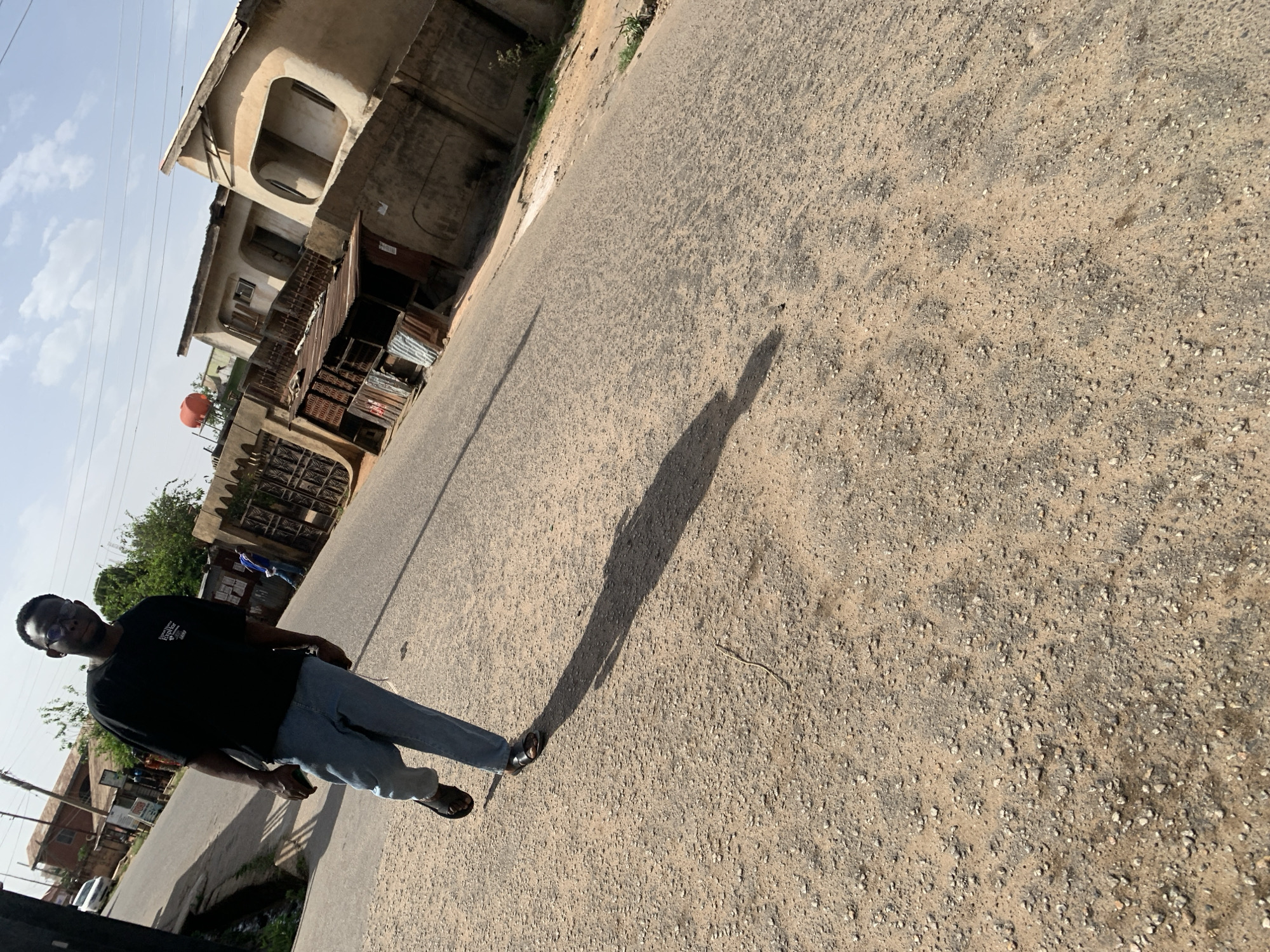 A full-length photograph depicts a man standing on a gravel road in Jos, Nigeria. The man is wearing a black T-shirt, blue jeans, and dark sandals. His shadow is cast on the road to his right. The road is made of compacted dirt and small stones. In the background, there are buildings, some with weathered exteriors. One building has a red water tank on its roof. Trees and greenery are visible along the roadside. Utility poles and wires are also present. The sky is visible with some clouds.
