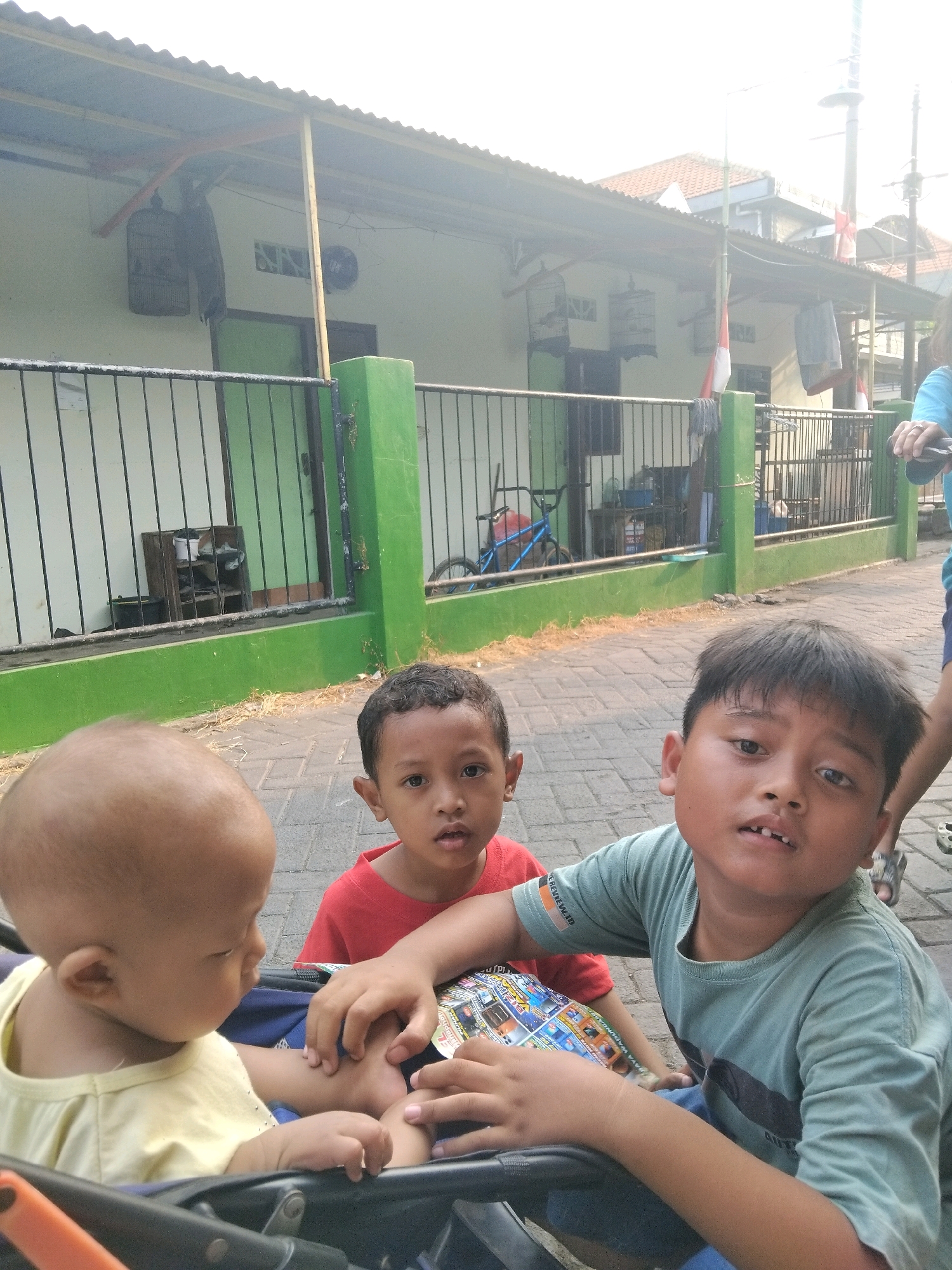 In the city of Kepuh Kiriman, Indonesia, three children are the subjects of this outdoor photograph. In the foreground, a baby with a shaved head, wearing a yellow shirt, is seated in a stroller. To the baby's right, a boy in a red t-shirt, with dark hair and brown eyes, looks towards the camera. Beside him, an older boy in a green t-shirt, also with dark hair and brown eyes, gazes upward. His left hand rests on the baby's leg. The children are positioned on a paved walkway lined with a green fence. Behind the fence, a building with a light-colored wall and a corrugated metal roof is visible, with birdcages hanging from the overhang. The background shows more buildings and some utility poles, suggesting a residential area. The lighting is bright, indicating daytime.