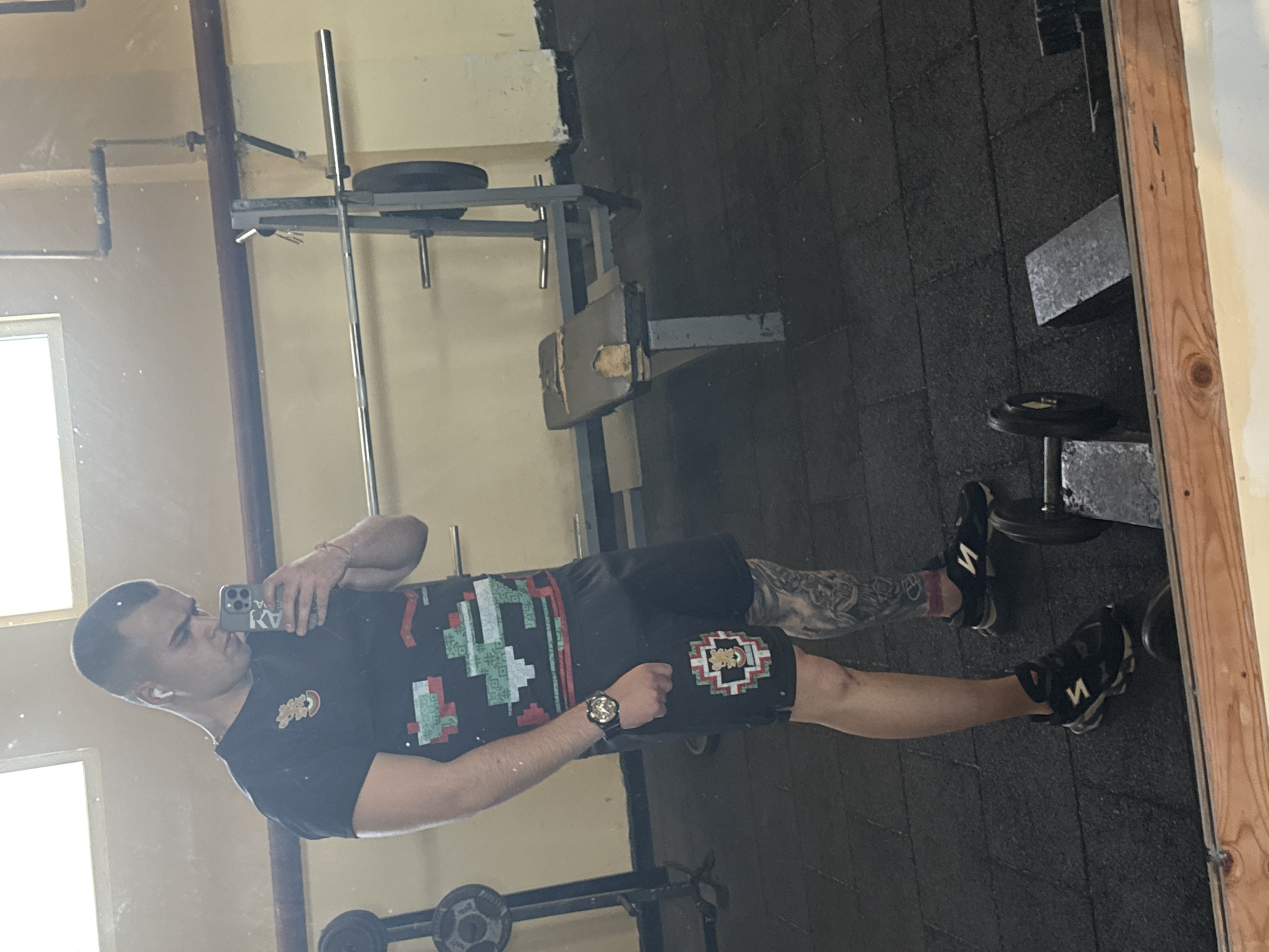 A medium indoor shot shows a man taking a selfie in front of a mirror in a gym in Unknown, Unknown. The man is wearing a black t-shirt with colored square patterns and black shorts with a matching pattern on the right leg. He has tattoos on his left arm and is wearing a watch on his left wrist. The background shows gym equipment, including a squat rack and dumbbells. The floor is black rubber. The mirror's wooden frame is visible on the right side of the image.