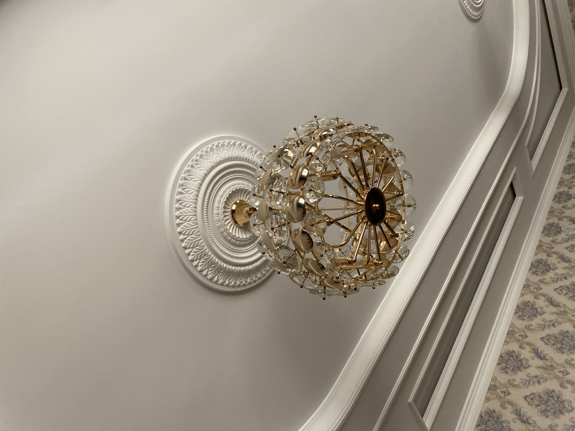 A photograph taken from a low angle looking up at a chandelier and ceiling details. The chandelier, made of gold-colored metal and clear crystal-like embellishments, hangs from a white decorative ceiling medallion. The ceiling itself has intricate white molding and decorative trim. In the lower portion of the image, a patterned wallpaper is visible. The scene appears to be an interior setting in a building. The provided information does not contain any details about the city of Turkistan, Kazakhstan, or any actions or people.