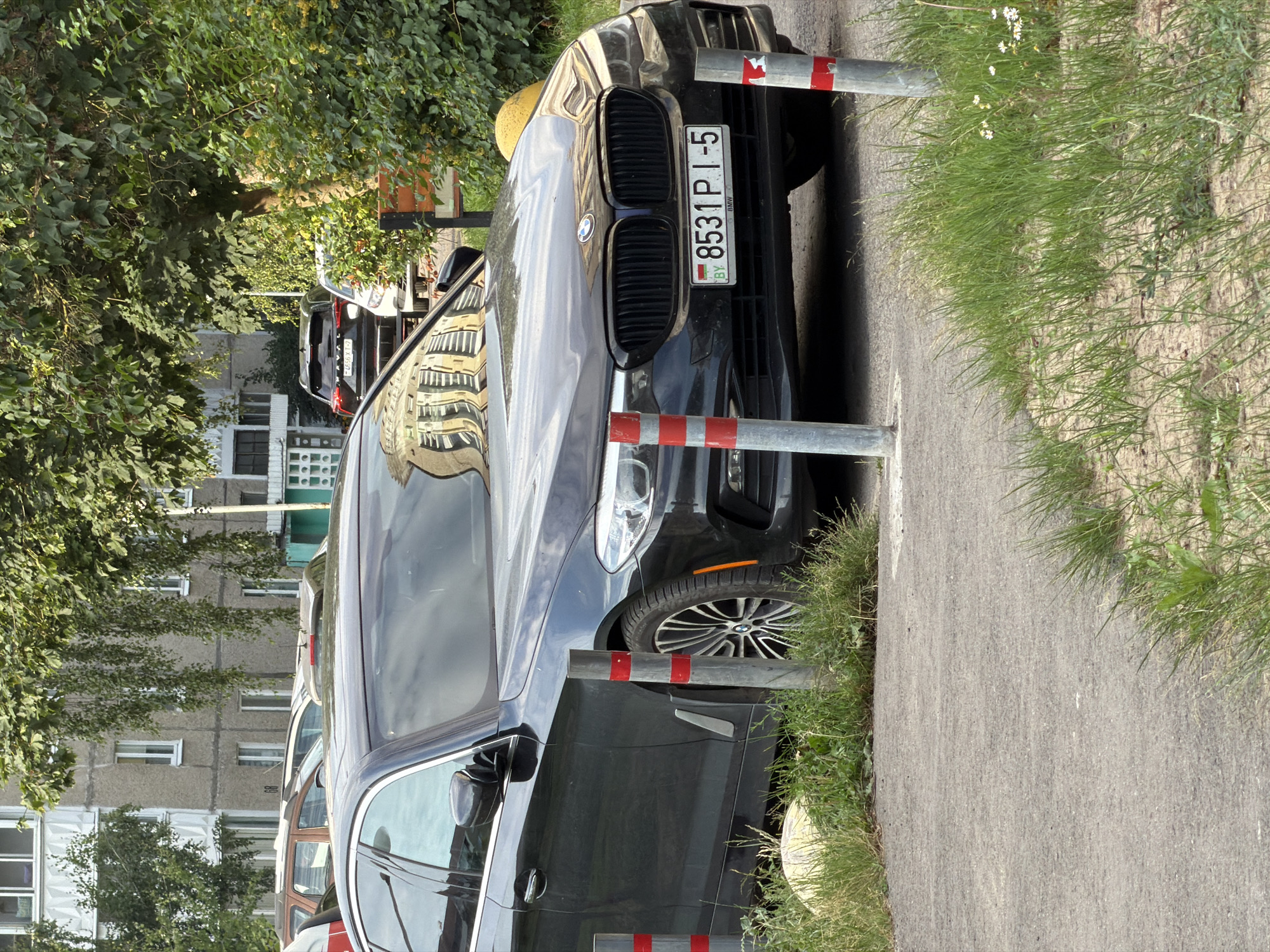 In Minsk, Belarus, a dark-colored sedan, identified as a BMW by its emblem and license plate, is parked on a paved surface adjacent to a grassy area. The license plate displays "8531 PI-5" and the country code "BY". The car's front grille, headlights, and part of its hood are visible. Two metal posts with red and white markings stand in front of the vehicle, partially obscuring it. In the background, a multi-story building with a beige facade and white window frames is partially visible, along with dense green foliage from trees. Another black SUV is parked further in the background.