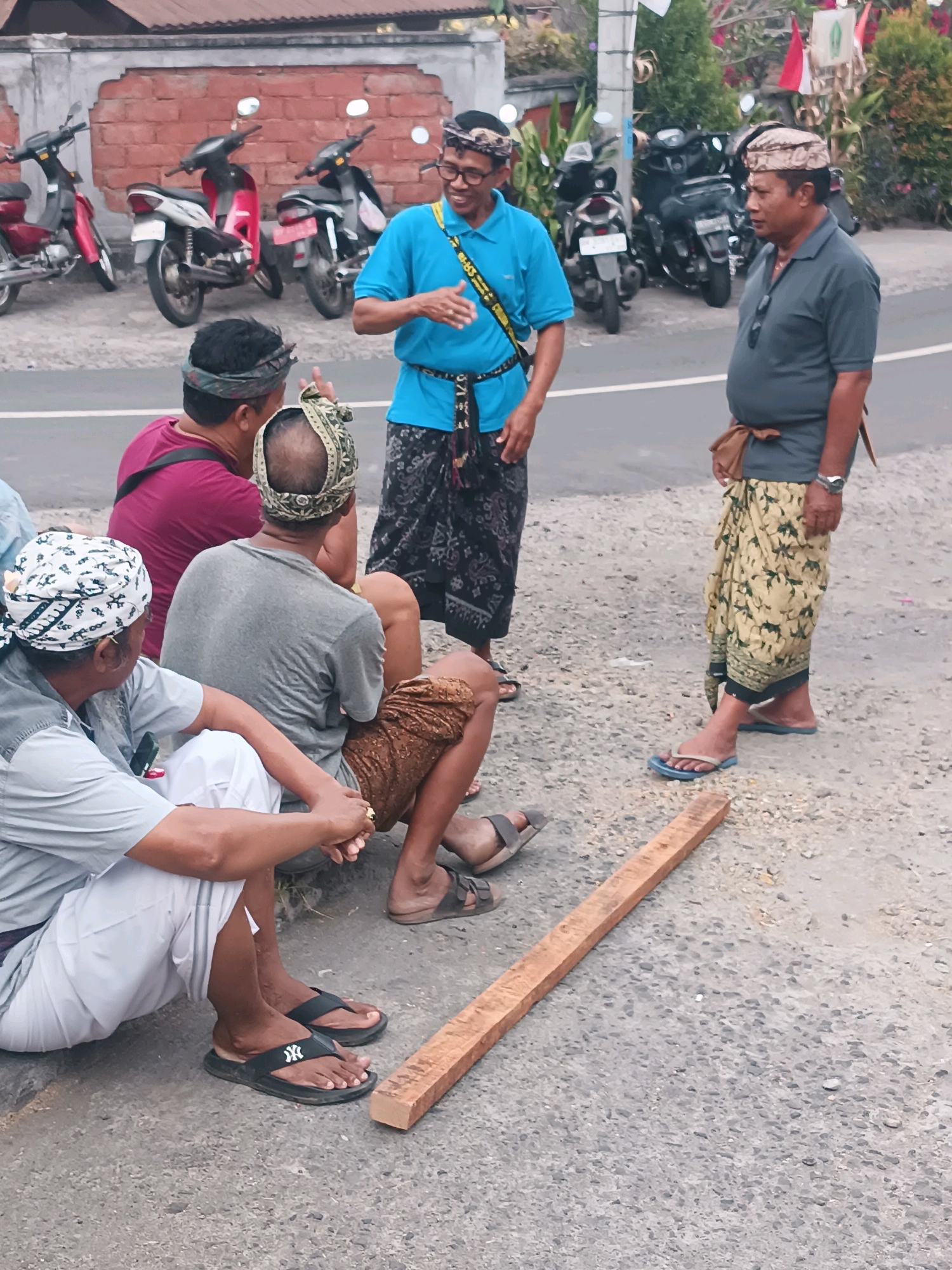 Four men are gathered on a paved surface with motorcycles parked behind them. One man, wearing a blue shirt and a patterned sarong, stands and gestures while speaking. Another man in a grey shirt and a patterned sarong stands opposite him, looking away. Two other men are seated on the ground. One wears a maroon shirt, and the other wears a grey shirt and white pants. A wooden plank lies on the ground between the standing men. The scene appears to be in Selemadeg, Indonesia, suggested by the traditional clothing and the presence of motorcycles.