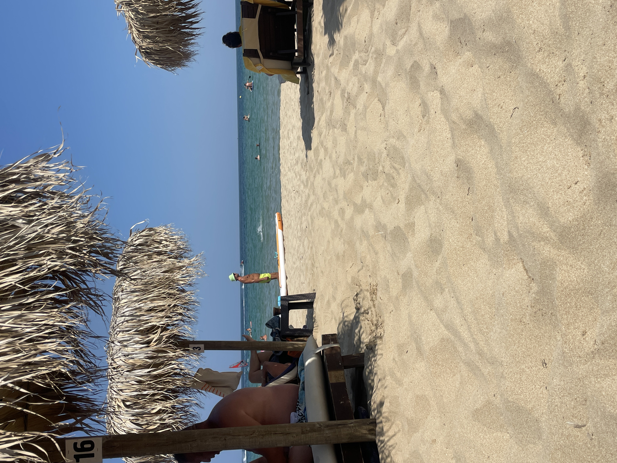 This is an outdoor, eye-level shot of a sandy beach with a clear blue sky. In the foreground, golden sand covers the ground, with some visible texture and shadows. Two beach umbrellas made of dried palm leaves are prominent, casting shadows on the sand. The umbrella on the left has a sign with the number "16" on its wooden pole.
Under the left umbrella, a man is partially visible from the chest up, facing left. He appears to be seated on a lounge chair. Nearby, a white towel with red stripes is hanging.
In the middle ground, the beach extends towards the sea. Several people are visible in the water, some swimming and others on paddleboards. There is a long, orange and white paddleboard lying on the sand near the water's edge. A dark-colored side table sits between two wooden posts of the umbrella, with a drink on it. Another person, a child wearing a green hat, stands on the sand near the water.
In the background, the turquoise sea stretches to the horizon. More people are in the water, and a few boats, possibly jet skis, are visible further out. The coastline is not clearly visible, but the water appears calm. On the right side of the image, another beach umbrella is partially visible, and a person is seated on a lounge chair facing the sea. The overall impression is a sunny, relaxing day at the beach.
While the exact location is specified as Primorsko, Bulgaria, this visual information is not directly discernible from the image content itself.