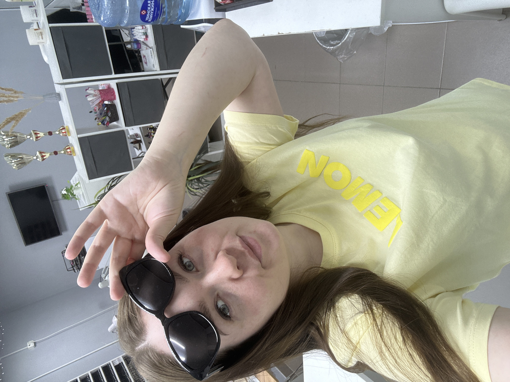A young woman is posing for a selfie in what appears to be an indoor setting. She is wearing a yellow t-shirt with the word "LEMON" printed on it, and sunglasses perched on her head. Her hair is long and brown. She is holding her hand up in a casual gesture.
The background shows elements of a room with a shelf unit filled with various items, including what looks like awards or trophies, and a large water bottle on top of the unit. A television is mounted on the wall. The wall appears to be light gray in color.
There is no information in the image to identify the exact location as the city of Chaikovsky, Russia.