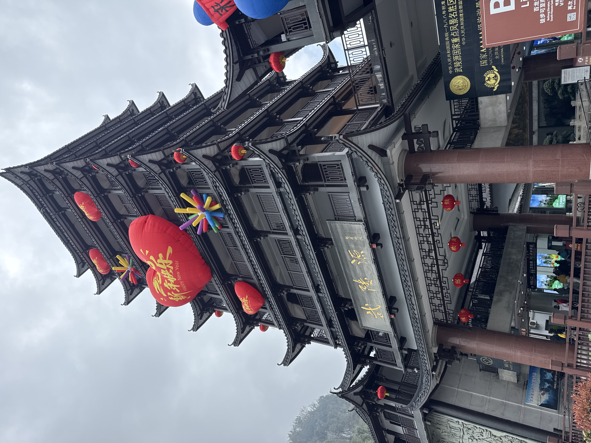 This image depicts the entrance to the Wulingyuan National Key Scenic Area in Wulingyuan, China, under an overcast sky. The prominent subject is a multi-tiered, traditional Chinese-style building featuring dark-tiled roofs, white walls, and intricate dark wooden balconies with railings. A large golden plaque bearing the characters "武陵源" (Wulingyuan) is mounted above the main entrance.
The building is extensively decorated with festive elements. Multiple large red heart-shaped inflatables are attached to the facade, one displaying "新年快乐 Happy New Year" in gold text. Several smaller red heart inflatables are also visible, along with colorful, spiky, starburst-like inflatables. Numerous traditional red Chinese lanterns are suspended from the eaves and connecting structures throughout the scene.
At ground level, the structure is supported by large, red-brown columns. Below the main plaque, a gold sign confirms the location as "中华人民共和国国务院一九八八年批准 武陵源国家重点风景名胜区" (Wulingyuan National Key Scenic Area approved by the State Council of the People's Republic of China in 1988) and a "国家AAAAA级旅游景区" (National AAAAA Tourist Attraction).
Approximately 10-15 individuals are visible at the ground-level entrance, appearing to queue or move through a designated entry point labeled "A入口" (A Entrance). Digital display screens are present at the entrance, one indicating "卫生间" (Restroom) and "饮水供应点" (Drinking water supply point). Another sign references the "百龙天梯" (Bailong Elevator). To the left, a grey concrete wall with relief carvings of mountains connects to the main building. A brown wooden fence runs along the bottom foreground, with some red-leaved foliage visible. Misty mountains are partially visible in the distant background to the left, obscured by the cloudy conditions. A skyway connects the main building to an adjacent structure on the right, which is also adorned with a blue inflatable and a red banner.
