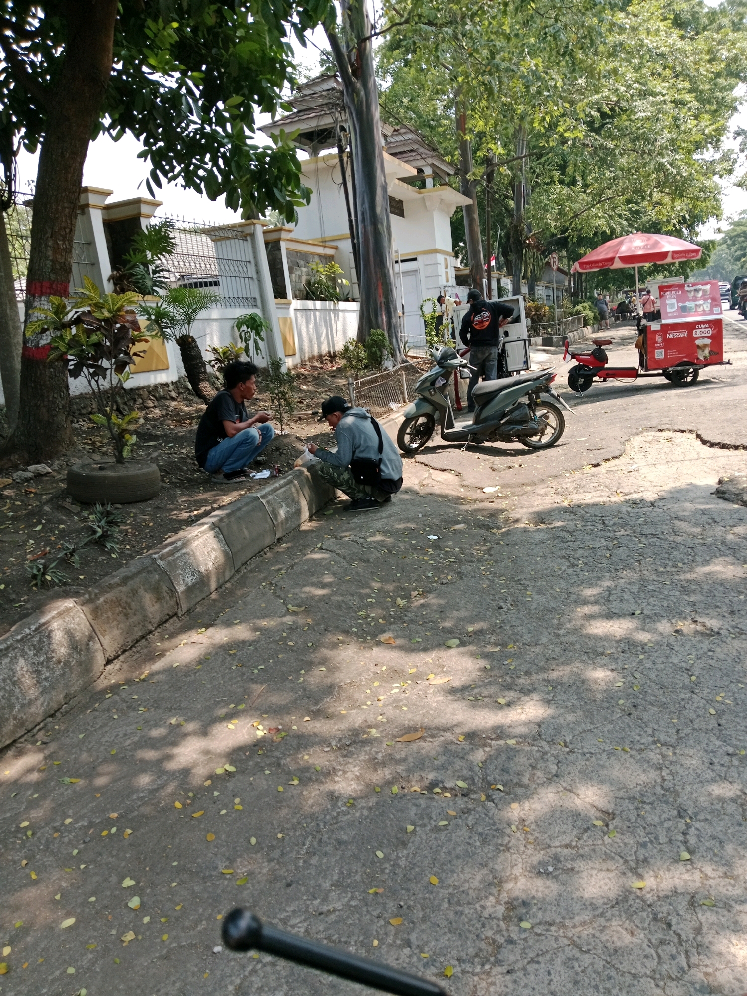 The image depicts a street scene in Karangtengah, Indonesia, on a bright, sunny day. In the foreground, a cracked asphalt road is covered with fallen leaves, with shadows from trees dappling the surface. To the left, a concrete curb separates the road from a grassy area. Two young men are seated on the ground near the curb. One, wearing a black t-shirt and blue jeans, is looking at his hands. The other, in a grey hoodie and black cap, is facing away from the camera, holding a plastic bag and appearing to interact with something within it. A motorcycle is parked on the road to the right of the men. Further down the street, a food cart with a red umbrella is visible. The cart is red and white and has advertising for Nescafe. A person is standing next to the cart, and another red motorized vehicle is attached to the cart. A white building with a white fence and decorative yellow accents is situated behind the grassy area on the left. The overall environment is that of a moderately busy street in a residential or commercial area.