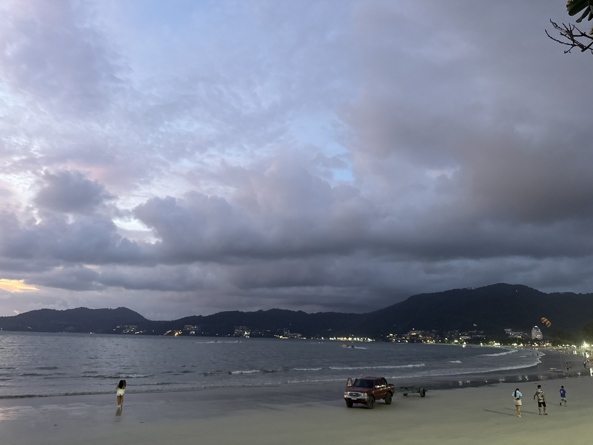 The image depicts a twilight scene on Patong Beach, Thailand. The sky is a canvas of dramatic, bruised clouds tinged with soft pink and blue, indicating either the very end of sunset or the approach of dawn. Dark, tree-covered hills form a backdrop to the bay, their slopes dotted with the twinkling lights of buildings. The sea is calm, with gentle waves lapping at the sandy shore.
On the beach, a solitary figure, appearing to be a young woman, stands at the water's edge, facing the sea. Further down the beach, a red SUV is parked with its driver's side door open. Beside it, a small trailer is visible. A few other individuals are scattered along the shoreline, some walking, others standing. In the distance, several boats are visible on the water, and further along the coast, more lights indicate the presence of businesses and resorts. A parasail can be seen in the air above the distant hills, adding a touch of activity to the otherwise tranquil scene. The overall atmosphere is one of quiet transition, as day gives way to night.