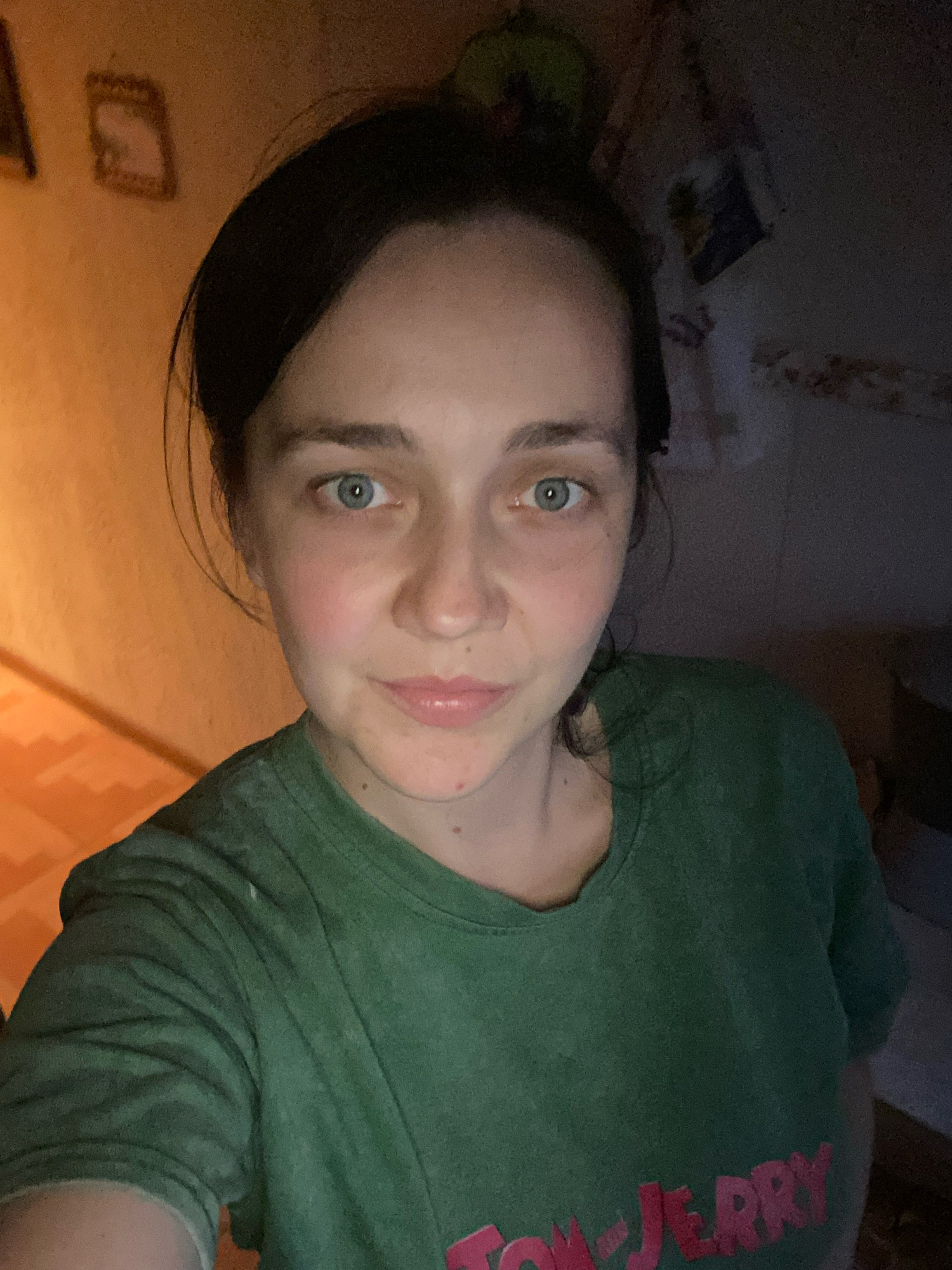 A close-up, high-angle selfie of a young Caucasian woman with dark hair and light blue eyes is shown. She is looking directly at the camera and wearing a green t-shirt with a red "TOM & JERRY" graphic. The background is dimly lit and appears to be a domestic indoor setting with a textured orange wall to the left and a dark, cluttered area to the right. The lighting suggests artificial light sources, with a warm glow on the left side of the image and a darker, more muted tone on the right. The overall impression is that of a casual, indoor photograph taken in a dimly lit environment.