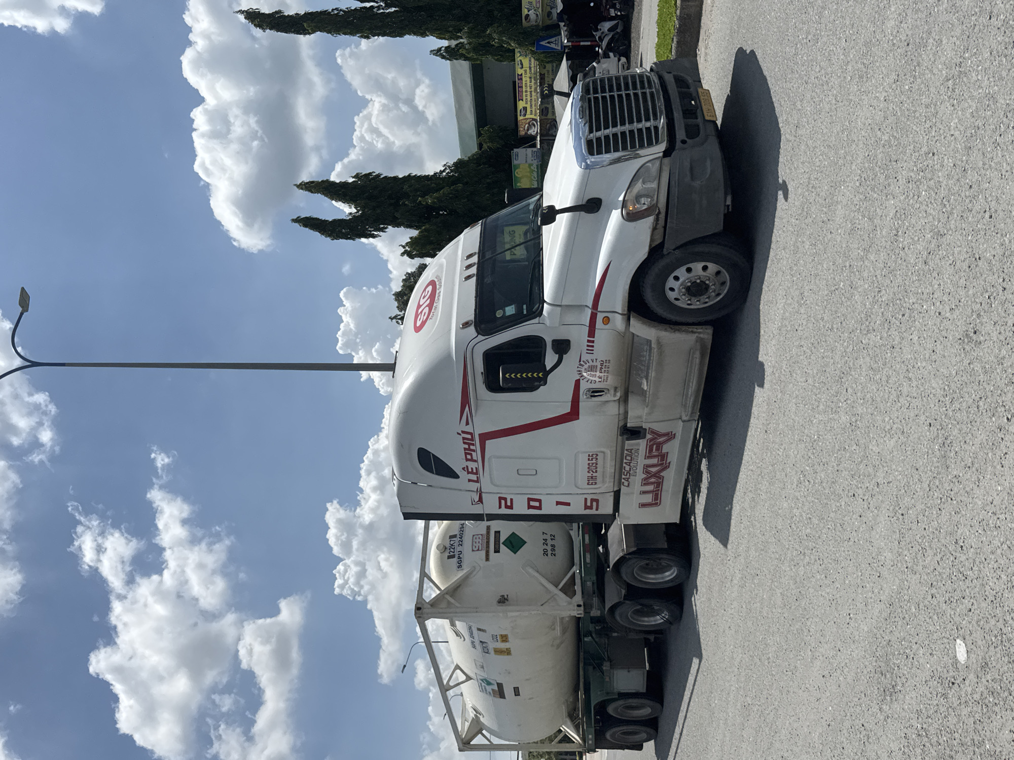 A white semi-trailer truck with a white tank trailer is parked on the side of a road in Ben Cau, Vietnam. The truck has the number "2015" printed on its side, and "LE PHU" and "LUXURY" are also visible. The trailer is carrying a large white cylindrical tank. In the background, there are trees and buildings. The sky is blue with scattered white clouds. The road appears to be asphalt.