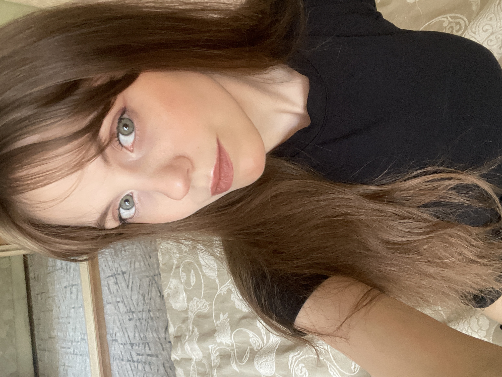 A selfie of a young woman is presented, taken from a slightly high angle. She has fair skin, light green eyes, and long brown hair with bangs. She is wearing a black top. The background appears to be a beige-patterned bedspread. The location cannot be determined based on the image alone, as there are no specific landmarks or identifying features to indicate Ufa, Russia, or any particular place.