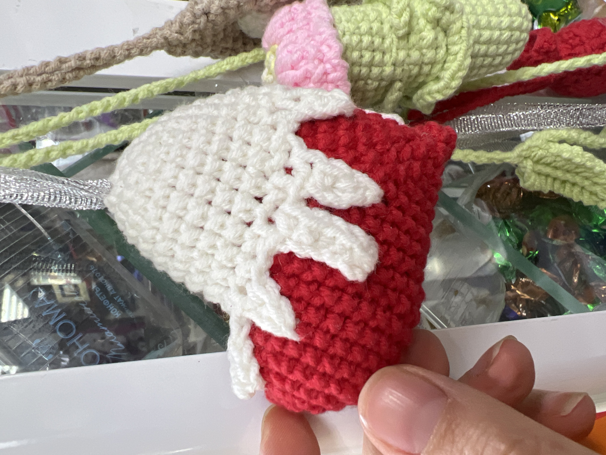 A person's hand holds a small, red crocheted item topped with white crocheted "icing" or "cream," resembling a festive cupcake or dessert, in an indoor setting in Chaikovsky, Russia. The hand is positioned towards the bottom right of the frame, with the fingers gently supporting the base of the item.
In the background, a variety of other colorful crocheted elements are visible, including light green strands resembling leaves or vines, a pink ruffled section that could be part of a flower or topping, and a beige/brown crocheted rope. Silver metallic ribbon or tinsel is also intertwined among these items. Beneath the crocheted elements, several shiny, crinkled wrappers, primarily in green and brown/gold, suggest the presence of wrapped candies or chocolates.
A clear plastic or glass surface lies beneath some of the items. Through this surface, text is visible, written in Cyrillic. Key words that can be deciphered include "OHOMA" (possibly part of a brand name), "КОНФЕТЫ" (KONFETI, meaning "candies" or "sweets"), "МАРМЕЛАД" (MARMELAD, meaning "marmalade" or "jelly candy"), and "АВГУСТ 016" (AVGUZT 016, indicating "August 2016"). This suggests the items are resting on or in a package of confectionery. The overall scene conveys a cheerful, possibly decorative, arrangement featuring handmade crafts and sweets.