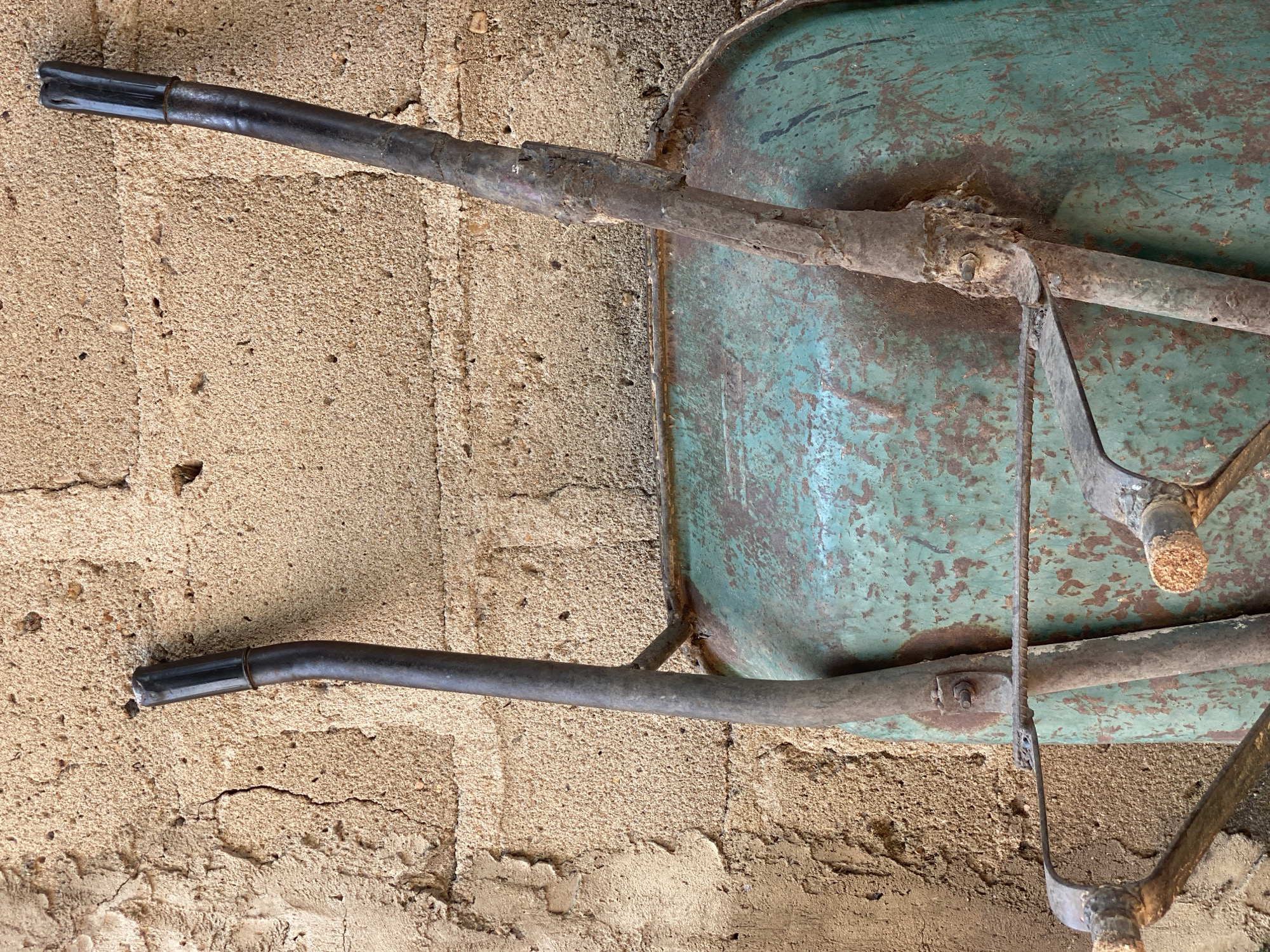The image shows the handles and top of a weathered, teal-colored wheelbarrow leaning against a textured, tan-colored wall. The wall appears to be made of concrete or stucco with visible cracks and an uneven surface. The wheelbarrow exhibits signs of rust and wear, particularly on its metal frame and handles. The handles are dark gray, with black plastic grips at the ends, and show some damage. A horizontal metal bar is visible across the top of the wheelbarrow. The scene appears to be an outdoor setting, possibly a yard or an alley, in a location consistent with Daniel, Nigeria, given the architectural style of the wall and the likely use of such a tool in that environment. No people or specific actions are depicted.