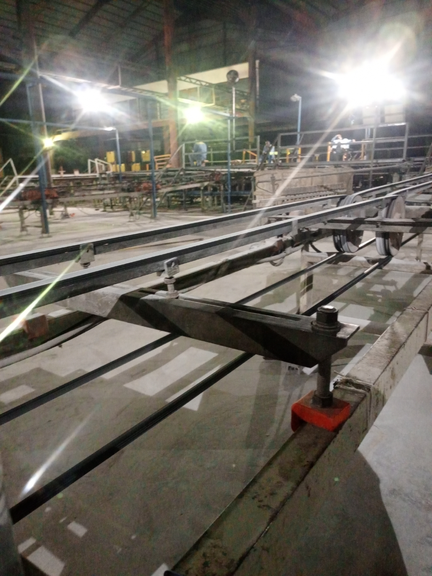 This image depicts an active industrial facility in Magboro, Nigeria, illuminated at night by multiple bright artificial lights causing significant lens flare. The foreground features a large, elongated metal conveyor system or track structure, comprised of parallel rails, support beams, and visible rollers/pulleys. Several clamps and fasteners secure parts of this system, including a distinct orange-colored clamp on a lower beam in the right mid-ground. The floor is a light-colored, flat surface, likely concrete, reflecting some light. In the background, a complex arrangement of industrial machinery, platforms, and scaffolding-like structures extends across the scene. Elevated platforms host several individuals: one person in light-colored attire (possibly a blue shirt) is visible on a platform to the left of a prominent light source, and another individual is seen leaning against a railing on a platform to the right of the same light. Additional indistinct figures are present further in the background. Yellow containers or equipment are discernible in the far background. The overall environment suggests a manufacturing or processing plant operating during non-daylight hours.