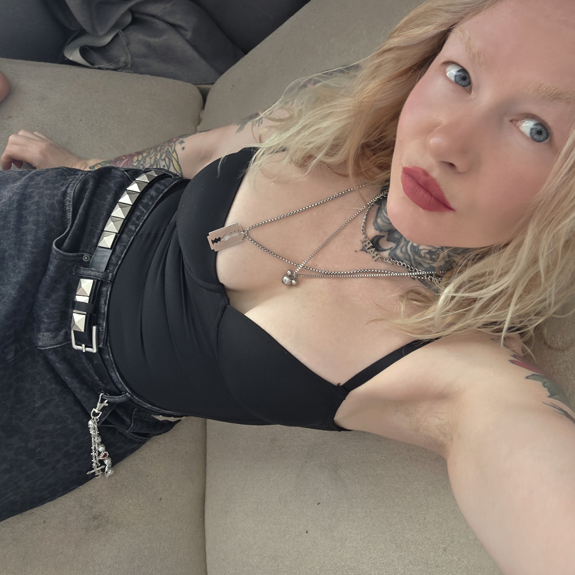 A woman is lying on a light-colored sofa, posing for a selfie. She is fair-skinned with blonde hair and blue eyes, wearing a black top, black jeans with a studded belt, and necklaces, one of which has a razor blade charm. She has tattoos on her neck and arms. The location is not Saint Petersburg, Russia.