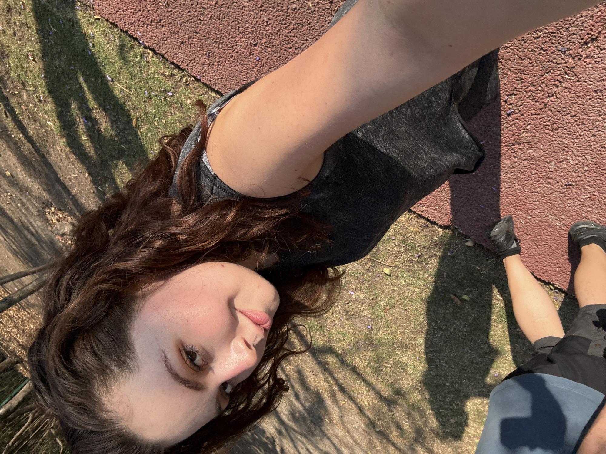 A selfie, taken in daylight, shows a fair-skinned person with curly brown hair wearing a gray tank top. The photo is angled downwards, capturing the person's face, shoulder, part of their torso, and a section of their legs and feet. The person is smiling slightly and looking at the camera. They are standing on a surface that transitions from a red, textured material to grass with scattered purple flowers. Tree shadows are visible on the grass. The location appears to be outdoors. There are no readily available details in the image to specifically identify the location as Santa Clara Ocoyucan, Mexico.