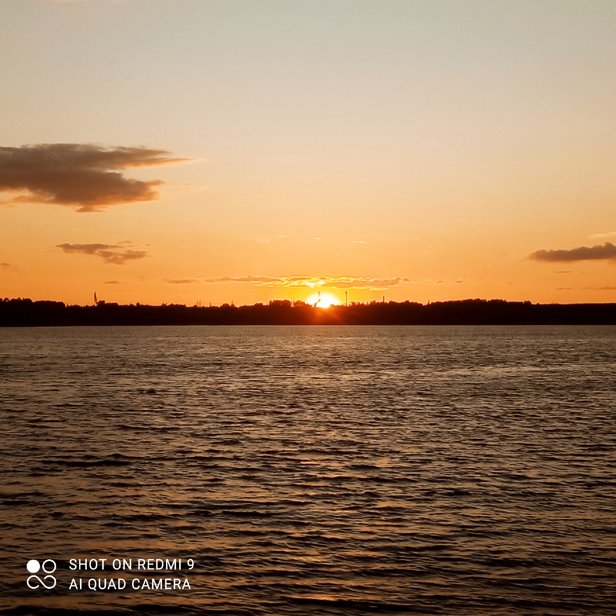 The image shows a wide body of water, likely the Kama River, in Chaikovsky, Russia, at sunset. The sky is filled with warm orange and yellow hues, transitioning to a lighter beige at the top, with scattered, elongated clouds. A dark silhouette of trees and industrial structures lines the horizon. The setting sun is partially obscured by a cloud, casting a bright glow on the sky. The water's surface is rippled, reflecting the colors of the sunset. In the bottom left corner, there is a watermark that reads "SHOT ON REDMI 9 AI QUAD CAMERA."