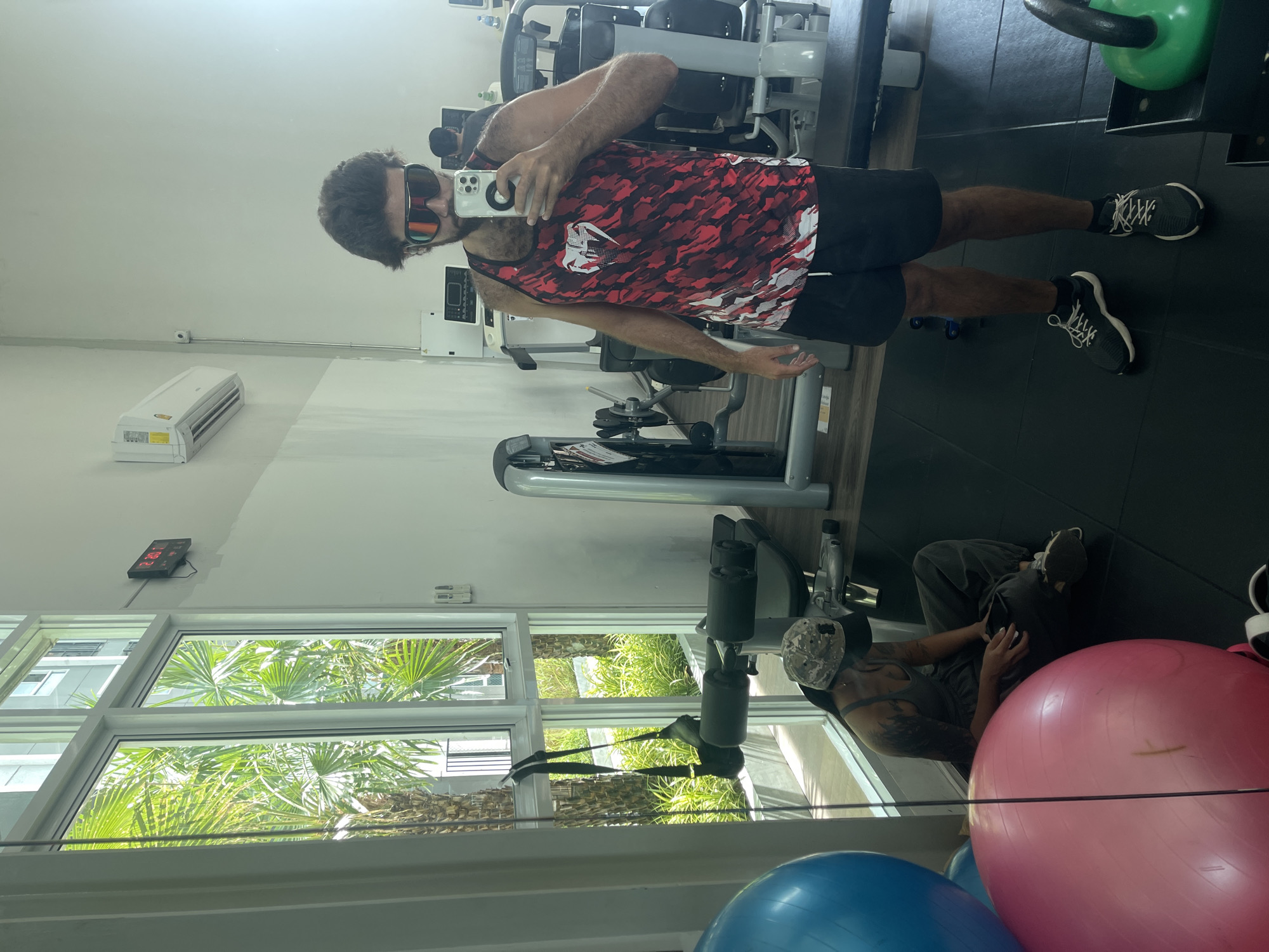In a brightly lit gym, a man with dark, tousled hair and a beard is taking a mirror selfie. He is wearing large, wraparound sunglasses, a red and black patterned tank top, and black shorts. He is holding a white smartphone up to take the photo. To his left, a person with tattoos on their arm is sitting on the floor next to two large exercise balls, one pink and one blue, and looking at their phone. The gym has large windows that look out onto lush green palm trees and a building, suggesting an urban or tropical setting like Bangkok, Thailand. Various gym equipment, including weight machines and a kettlebell, are visible in the background. A digital clock on the wall shows "12:30". An air conditioning unit is mounted high on the wall.