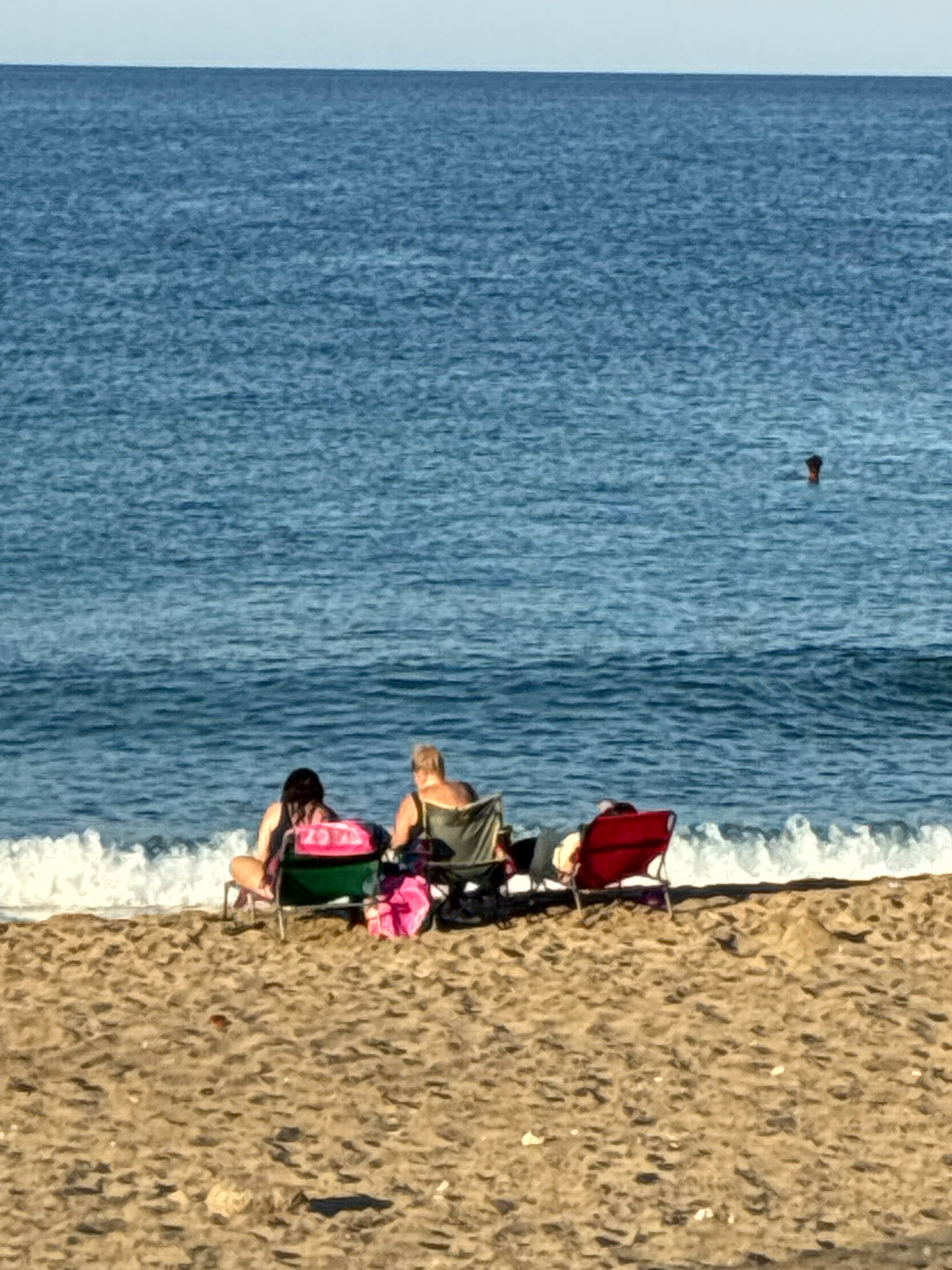 The image shows a beach scene on a sunny day with a calm sea in the background. Three people are visible sitting on beach chairs on the sandy shore. In the foreground, two individuals are seated on chairs. The person on the left is facing away from the camera and is wearing dark clothing. They are sitting on a green beach chair next to a pink bag. To their right, another person is seated on a similar chair, facing left, and is wearing a tank top. To the right of this person, a red beach chair is occupied by a person whose upper body is visible. In the middle distance, a person's head and shoulders are visible in the blue water, appearing to be swimming or floating. The sea stretches out to the horizon, meeting a pale sky. The sand is a light brown color, with some small debris scattered on it. The waves are gently breaking on the shore. The image appears to be taken in Alanya, Turkey, given the style of beach and the typical weather conditions.
