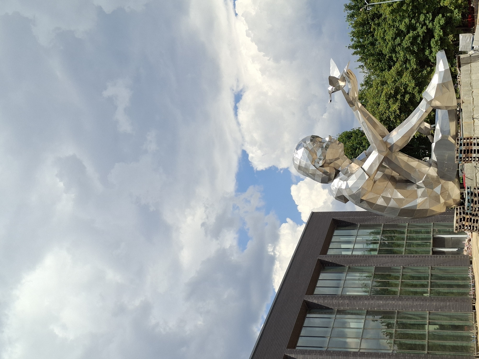 This is a photograph taken outdoors in Chișinău, Moldova, showcasing a large, faceted metal sculpture of a person holding a bird. The sculpture is seated and appears to be made of polished, reflective metal, possibly stainless steel or aluminum, with geometric planes. It is positioned in front of a modern building with a dark brick facade and large glass windows, reflecting the surrounding environment. Behind the sculpture and building, a dense canopy of green trees is visible under a partially cloudy sky. The overall scene suggests an urban park or public square setting. The sculpture itself is the dominant subject, its metallic form catching the light and creating a striking contrast with the natural elements and the building. The sky is filled with dramatic clouds, some appearing white and puffy, while others are a darker gray, suggesting a dynamic weather condition. The base of the sculpture is concrete and surrounded by what appear to be construction materials or pallets, indicating it might be a recent installation or undergoing maintenance. The photograph is taken from a slightly low angle, emphasizing the scale of the sculpture.
