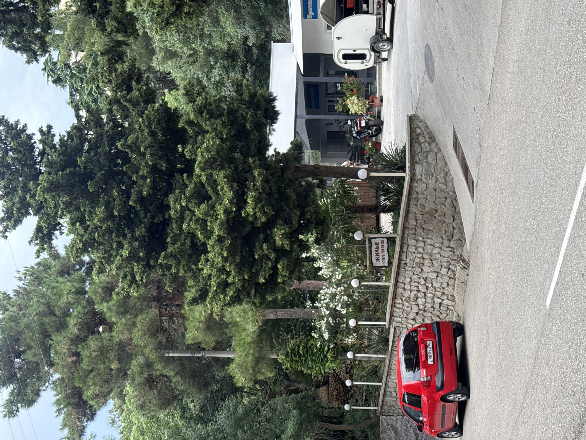 This is a street-level, eye-level, full-shot image of a residential or commercial area with lush greenery, likely in the city of Partenit, Ukraine. A bright red Hyundai Getz is parked on the asphalt road on the left side of the image. Behind the car, a stone wall separates the road from a manicured garden area with white spherical lights on poles and flowering white bushes. A sign with Cyrillic text, reading "ЖИЛЬЕ" and a phone number, is attached to a wooden post.
In the background, a modern building with a white roof and columns is visible, suggesting it could be a cafe, reception area, or shop. A motorcycle is parked in front of the building. To the right, a small, white teardrop trailer is hitched to an unseen vehicle, with its back door open, revealing some contents. The environment is dominated by mature trees, indicating a well-established, possibly resort-like, setting. The sky is overcast.