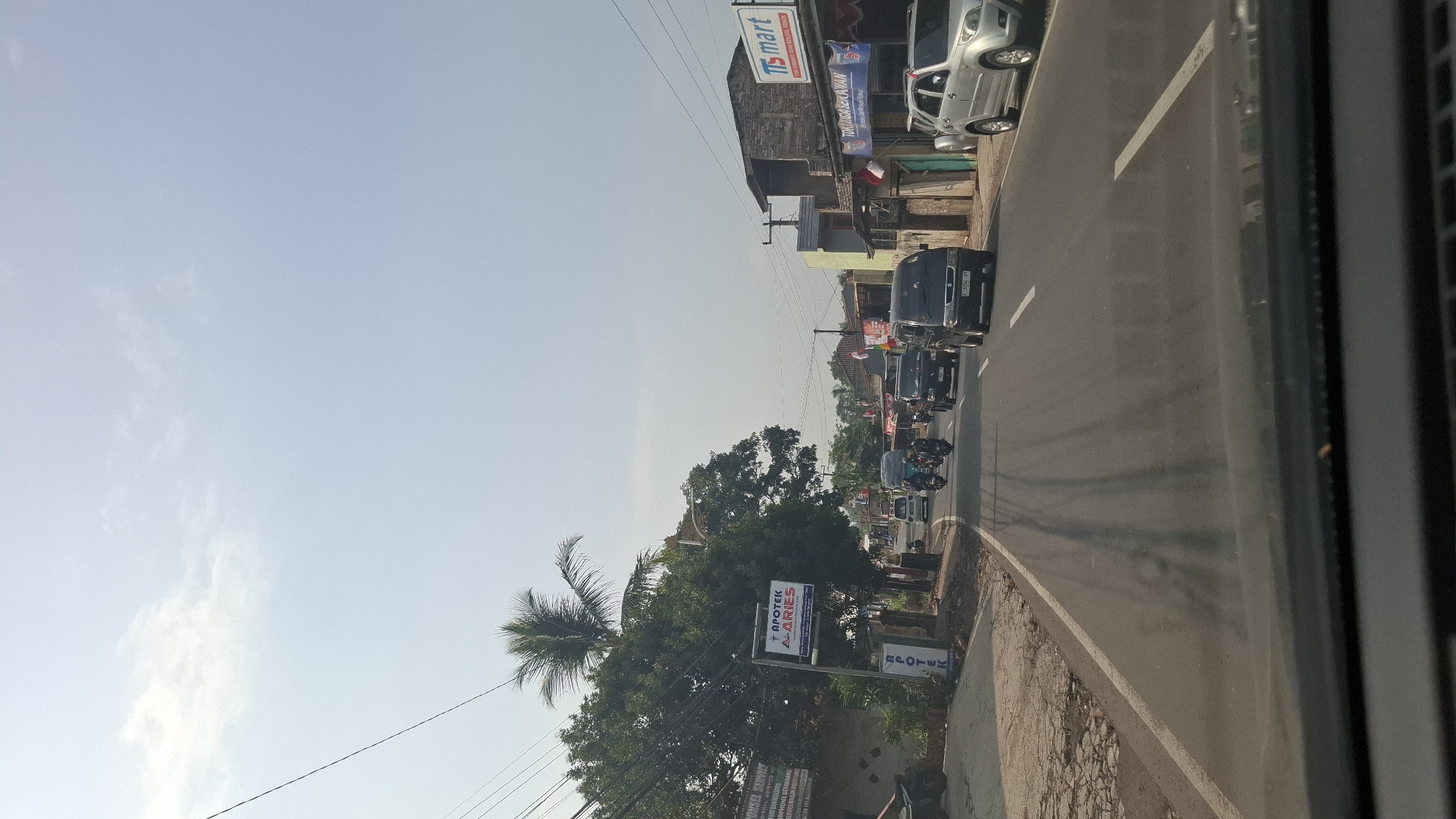The image depicts a street scene in Cilaku, Indonesia, likely during the daytime under a clear sky. The perspective is from inside a vehicle, looking forward. On the left side of the road, there are trees, a sidewalk, and a building with signage that includes "APOTEK ARIES" and "CONSEL SERVICE". Further down the road, there are several vehicles, including motorcycles and cars, traveling in the same direction. On the right side of the road, there are buildings with shops, including one with "TIS mart" signage and another with "TOKO DJAGA SEKAWAN" signage. Indonesian flags are visible, suggesting a national holiday or celebration. The road itself is paved with white lane markings. The overall environment appears to be a moderately busy semi-urban or suburban area.