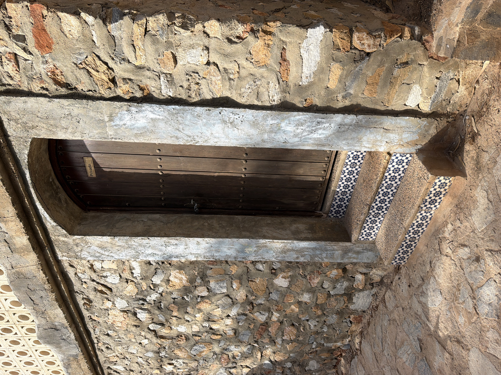The image shows an arched wooden door set into a stone wall. The door is flanked by a stucco frame that has some peeling paint. There are three steps leading up to the door, each decorated with blue and white geometric tiles. The wall itself is constructed from irregular stones of various colors, including brown, beige, and gray. Above the stone wall, there is a section of wall with a decorative pattern of circles. The ground in front of the door is paved with irregular stones. A small sign on the door reads "Privado". The scene appears to be outdoors in daylight.