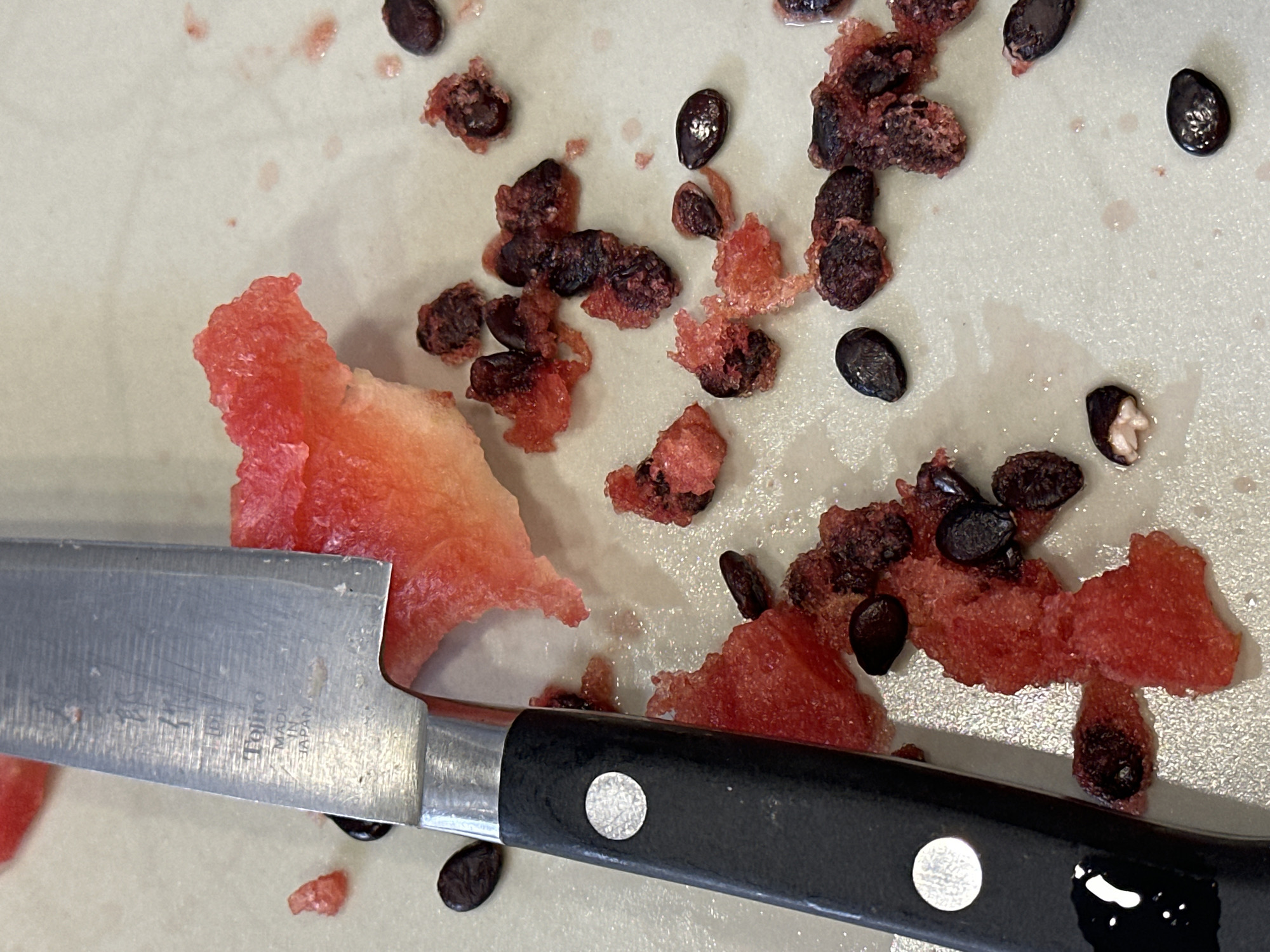 A close-up, overhead shot captures a kitchen scene in Nevinnomyssk, Russia, with a knife and watermelon remnants on a light-colored countertop. The knife, positioned on the left, features a silver blade with the brand "Tojiro" and "Made in Japan" etched into it, alongside a black handle. Scattered around the blade are pieces of red watermelon flesh and numerous dark watermelon seeds. Some seeds are partially embedded in the fruit, while others lie isolated. The countertop appears slightly wet, with a few small red smudges suggesting juice spills. The lighting is bright, highlighting the textures of the fruit and the metallic sheen of the knife.