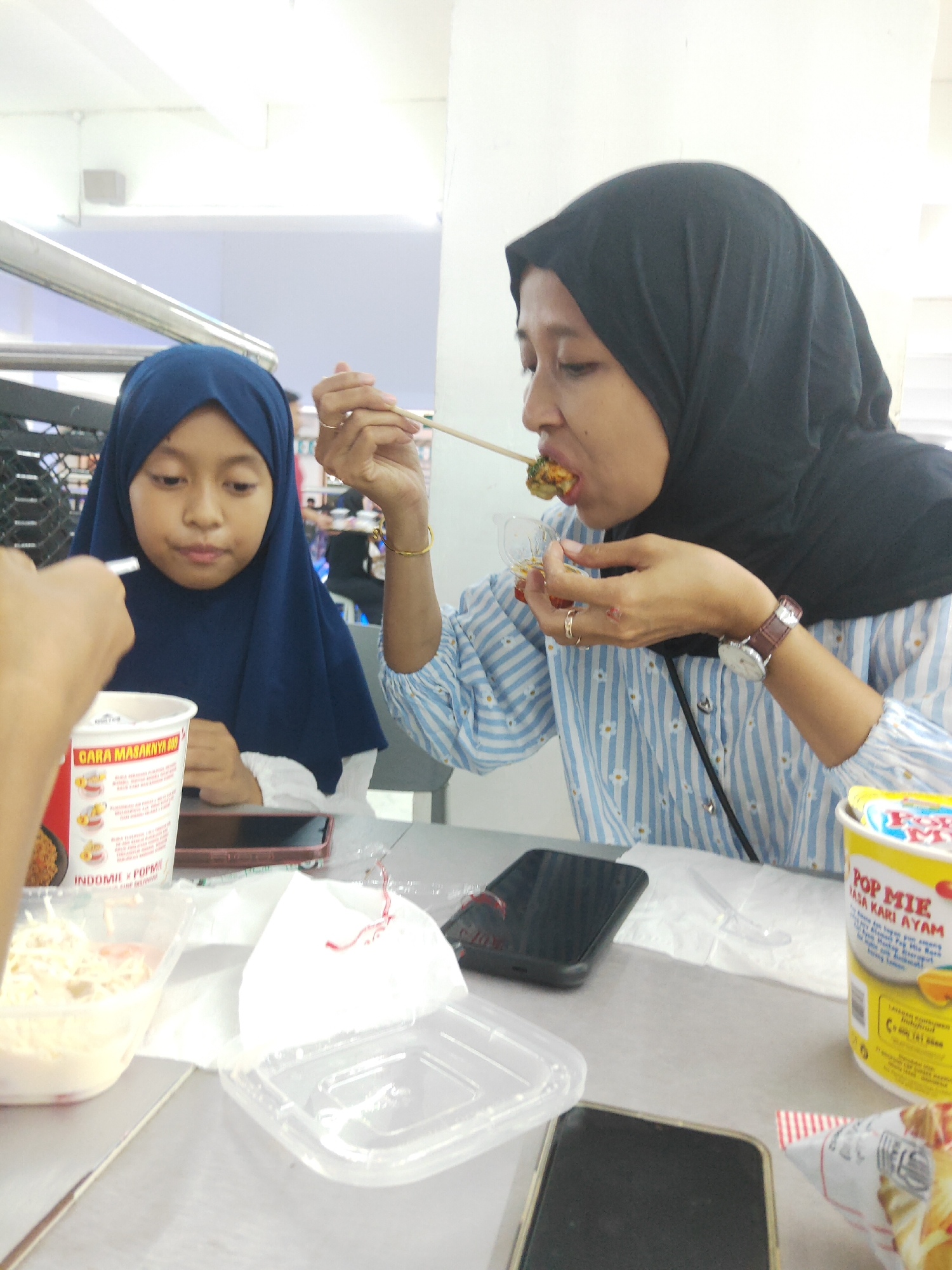 In Sintang, Indonesia, a young girl wearing a dark blue hijab sits at a table, looking down at a cup of instant noodles. Beside her, an adult woman wearing a black hijab and a blue and white striped shirt eats food with chopsticks. The table is cluttered with food containers, napkins, and cell phones. The setting appears to be a casual dining establishment, possibly a food court.
