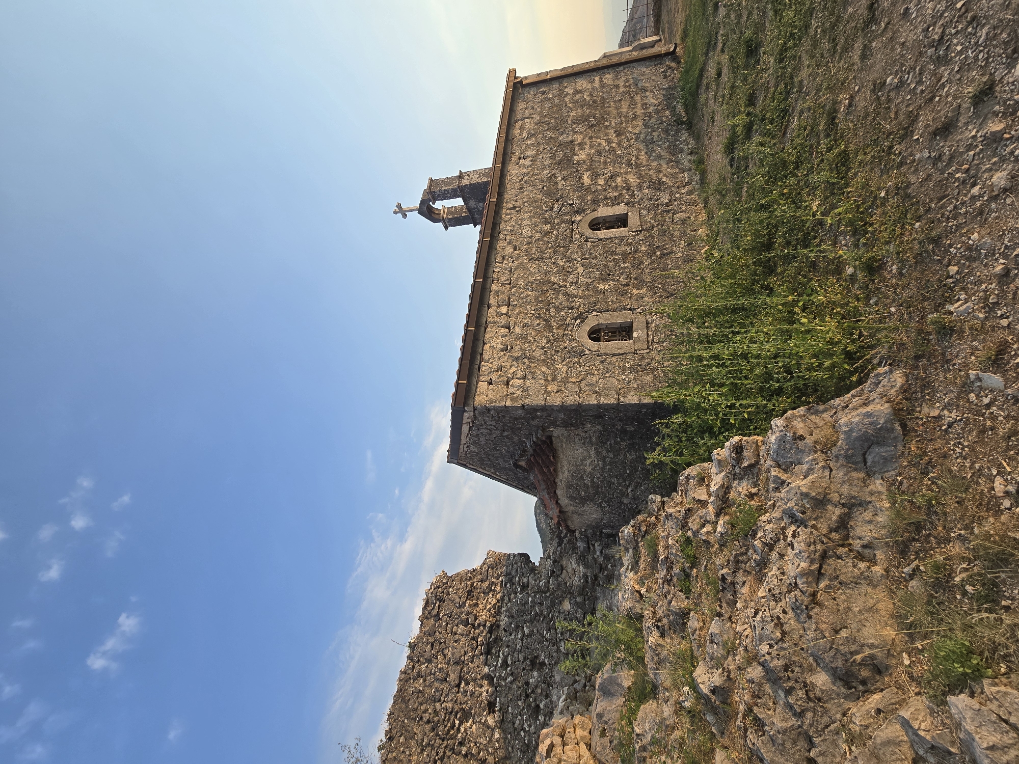 The image shows a small stone church with a bell tower topped with a cross. The church is located in Medun, Montenegro, amidst rocky terrain with scattered green vegetation. To the left of the church are the ruins of a stone structure. The sky above is clear blue with a few wispy clouds. The lighting suggests it is either sunrise or sunset. There are no people or animals visible in the image.