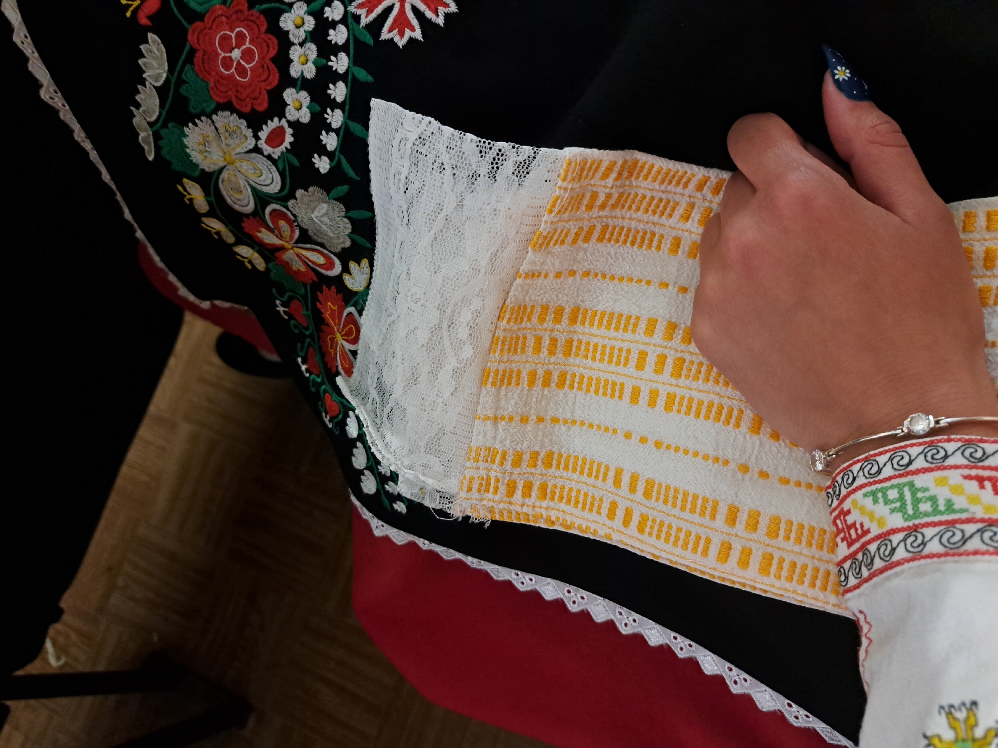 A close-up shot of a person wearing traditional Bulgarian clothing, potentially in Lokorsko, Bulgaria. The clothing consists of a black garment embroidered with colorful floral patterns, white lace detailing, a red layer with white scalloped trim, and a white sleeve with intricate, multicolored embroidery. The person's hand is visible, adorned with a bracelet and a long, dark blue fingernail decorated with a white daisy. They are holding a white fabric with yellow stripe patterns. The setting appears to be indoors, possibly a home or a cultural center.