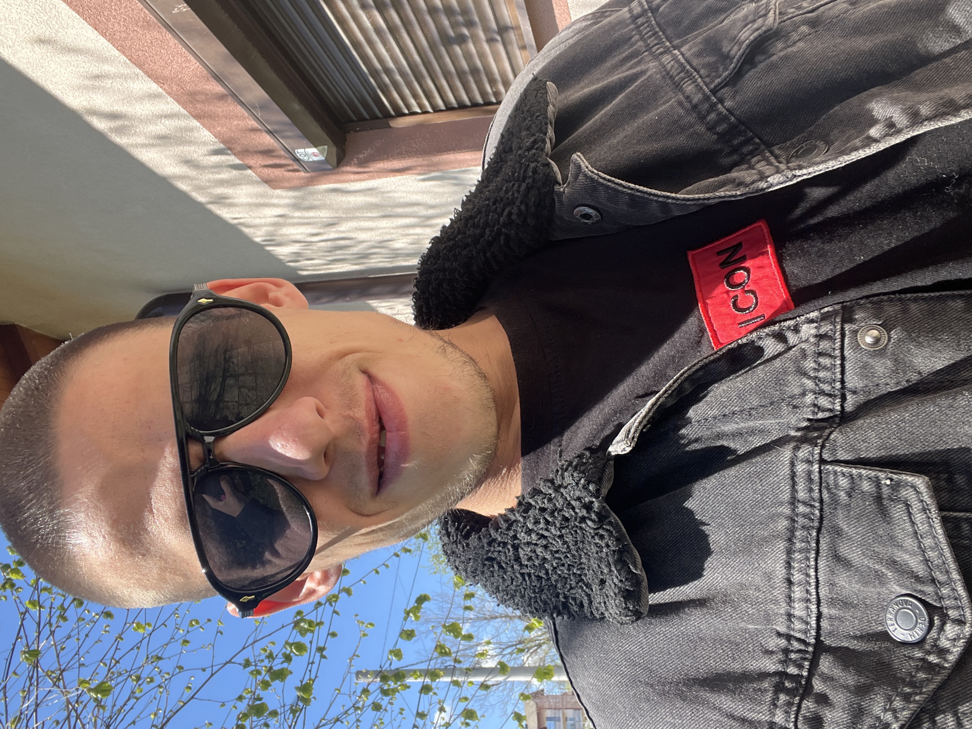 A man wearing sunglasses and a black denim jacket with a black shearling collar is facing the camera. The jacket has a red tag with "ICON" embroidered on it. The man has a short haircut and stubble. Behind him, there are trees with budding leaves and a blue sky. A building with a brown and white exterior is partially visible in the background.
The location appears to be in the city of Sofia, Bulgaria based on visual details.