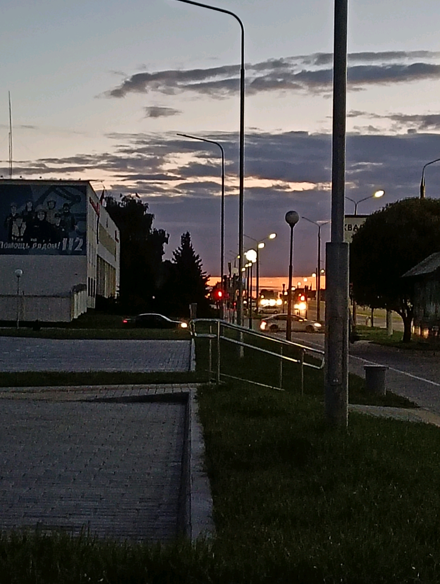 The image captures a street scene in Hrodna, Belarus, at dusk. The sky is filled with dramatic clouds, painted in shades of grey, purple, and orange from the setting sun. The foreground features a paved area with grass bordering it, and a metal railing runs along the edge of a slightly elevated pathway.
To the left, a building with a mural is visible. The mural depicts a group of people, and text in Russian that reads "Помощь рядом! 112" which translates to "Help is nearby! 112". A street lamp stands in front of the building.
The street itself is lined with multiple streetlights, some illuminated, casting a warm glow. Several cars are present on the road; one car in the mid-ground has its headlights on and appears to be moving. In the distance, more vehicles and lights suggest the presence of ongoing traffic. On the right side of the image, a portion of a building and trees are silhouetted against the twilight sky. The overall atmosphere is one of an urban environment transitioning into night.