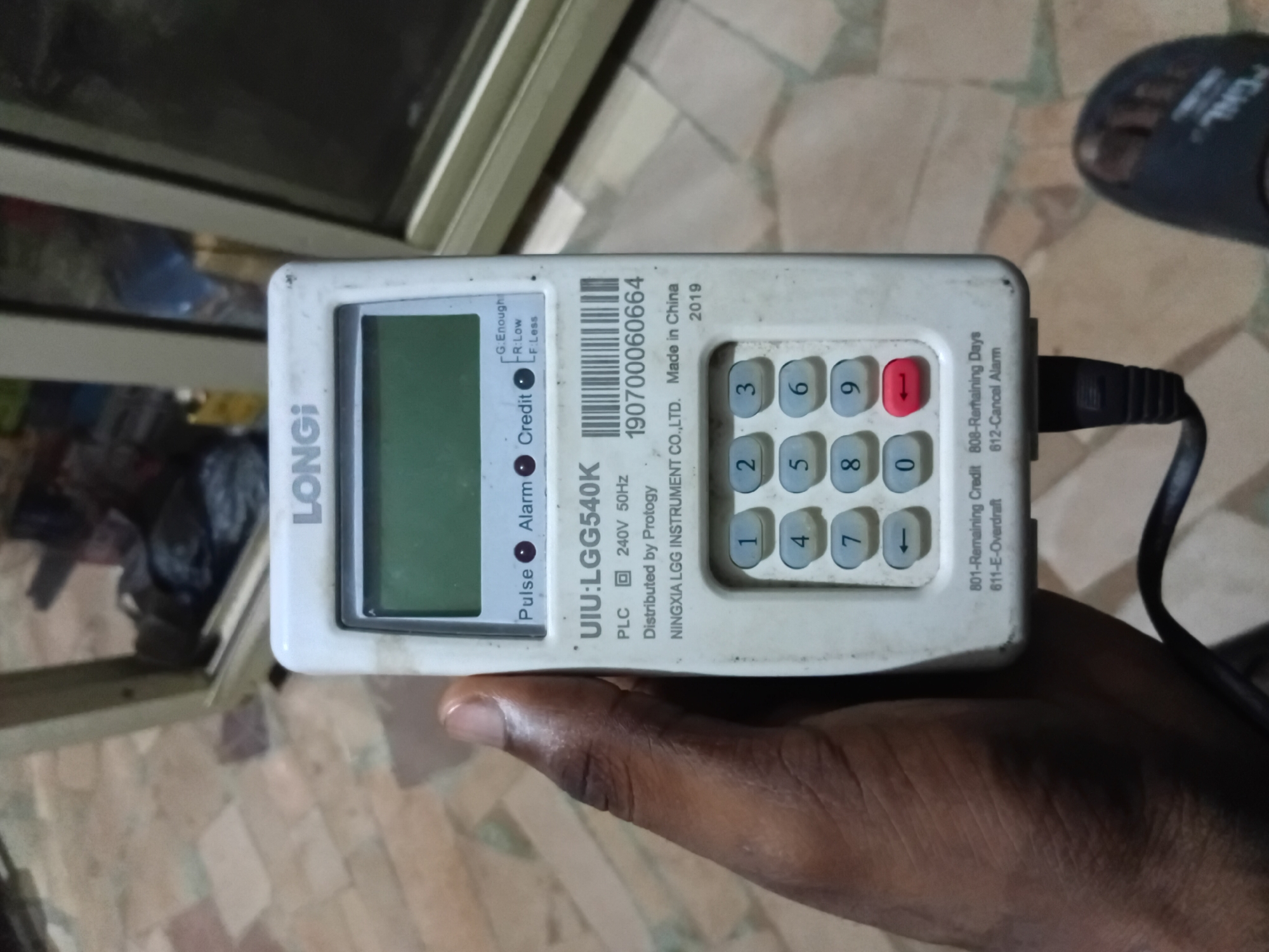 A hand holds a white LGG540K smart electricity meter in Nkpor, Nigeria. The meter has a digital display showing "LONGi" at the top. Below the display, there are three indicator lights labeled "Pulse," "Alarm," and "Credit." The display also shows text related to credit and alarm statuses. The meter features a numerical keypad with buttons labeled 0 through 9, along with arrow and red cancel buttons. Text on the meter indicates it is a UIU:LGG540K, PLC 240V 50Hz, distributed by Protogy, and manufactured by NINGXIA LGG INSTRUMENT CO.,LTD. Made in China, 2019. Below the keypad are codes for "Remaining Credit" and "E-Overdraft." The background shows a tiled floor and a blurry view of an indoor environment with some objects and reflective surfaces, likely within a building.