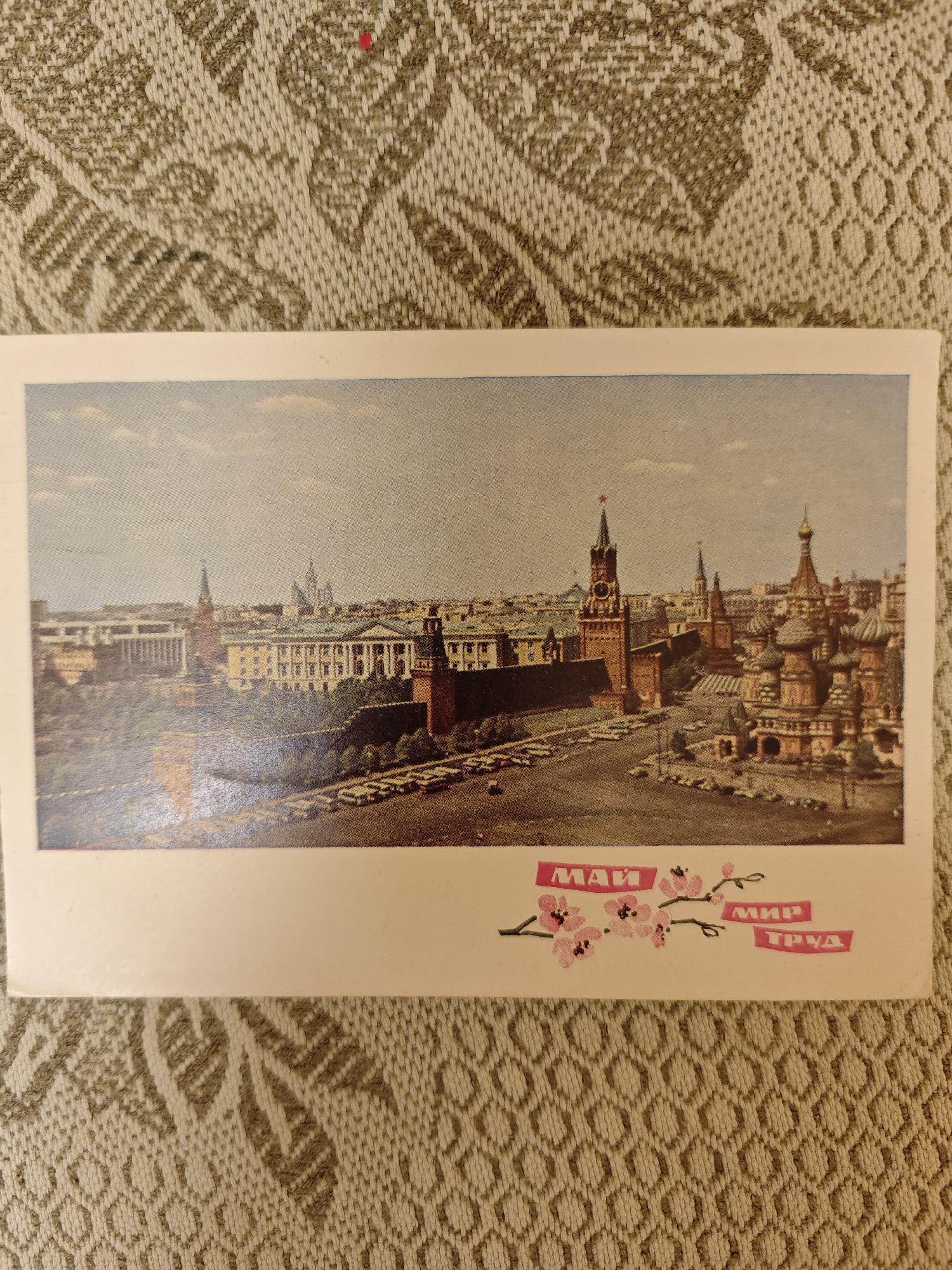 This image is a photograph of an old postcard, featuring an elevated view of Moscow, Russia. The postcard displays a wide cityscape dominated by iconic landmarks around Red Square. On the left, the distinctive red brick walls and several towers of the Moscow Kremlin are prominent, including the Spasskaya Tower with its clock face. To the right, the multi-colored, onion-domed St. Basil's Cathedral stands out against the sky. In the background, various other city buildings extend into the distance. Below the Kremlin walls, streets with parked cars and some trees indicate an active urban environment. The sky is a clear light blue with a few wispy clouds, suggesting a pleasant daytime setting.
The postcard's lower white border features decorative elements and text. In pink Cyrillic letters, the words "МАЙ" (May), "МИР" (Peace), and "ТРУД" (Labor) are displayed. These words are surrounded by delicate pink cherry blossom branches, a common motif. The combination of these specific words with the imagery of cherry blossoms strongly suggests the postcard commemorates May Day (International Workers' Day), a major celebration during the Soviet era. The style of the photograph and the vehicles visible indicate a likely production period from the mid to late 20th century.