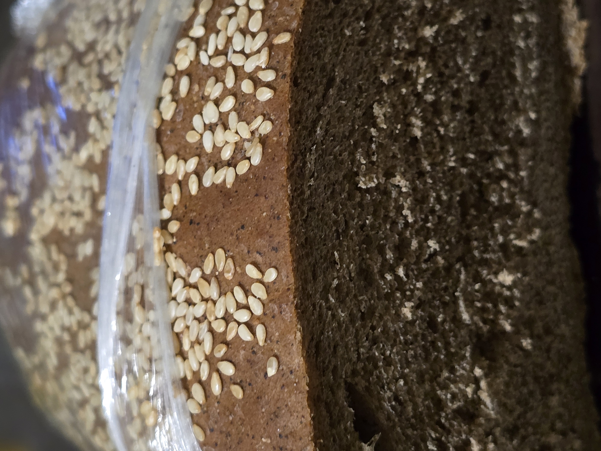 The image is a close-up, vertical shot of a loaf of bread, likely rye bread, covered in a plastic wrapper. The top of the loaf is visible, sprinkled with sesame seeds. The bread has a dark brown crust and a lighter brown, coarse interior texture. The focus is on the bread itself, with the background out of focus. There is no discernible information about the location in Nova Kakhovka, Ukraine.