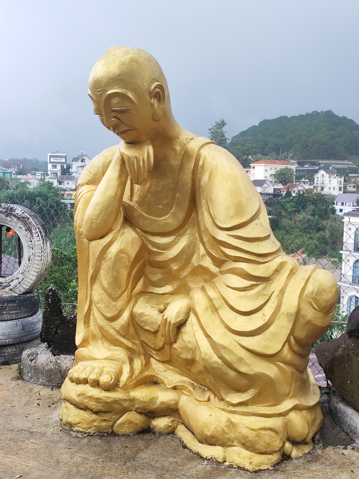 A golden statue of a contemplative figure, possibly an Arhat or a meditating Buddha, dominates the foreground and mid-ground of the image. The statue is depicted sitting with one hand gently supporting its chin, eyes cast downwards, conveying a pensive or peaceful expression. Its robes are intricately draped, showing folds and textures, all rendered in a bright golden hue that appears to be painted on concrete or a similar material. The statue's feet are visible at its base.
To the immediate left of the statue, a stack of worn, discolored tires is visible on the concrete ground. A chain-link fence runs horizontally behind the statue, separating the immediate area from the distant landscape. Beyond the fence, a sprawling town or residential area is visible, with numerous houses featuring various architectural styles, including white facades and tiled roofs, nestled among green trees and vegetation. In the far background, verdant, forested mountains rise against an overcast, light grey sky, suggesting a cloudy day.
The scene depicts a serene and reflective moment, likely situated on a high point overlooking the city of Da Lat, Vietnam, given the context. There is no visible text in the image.